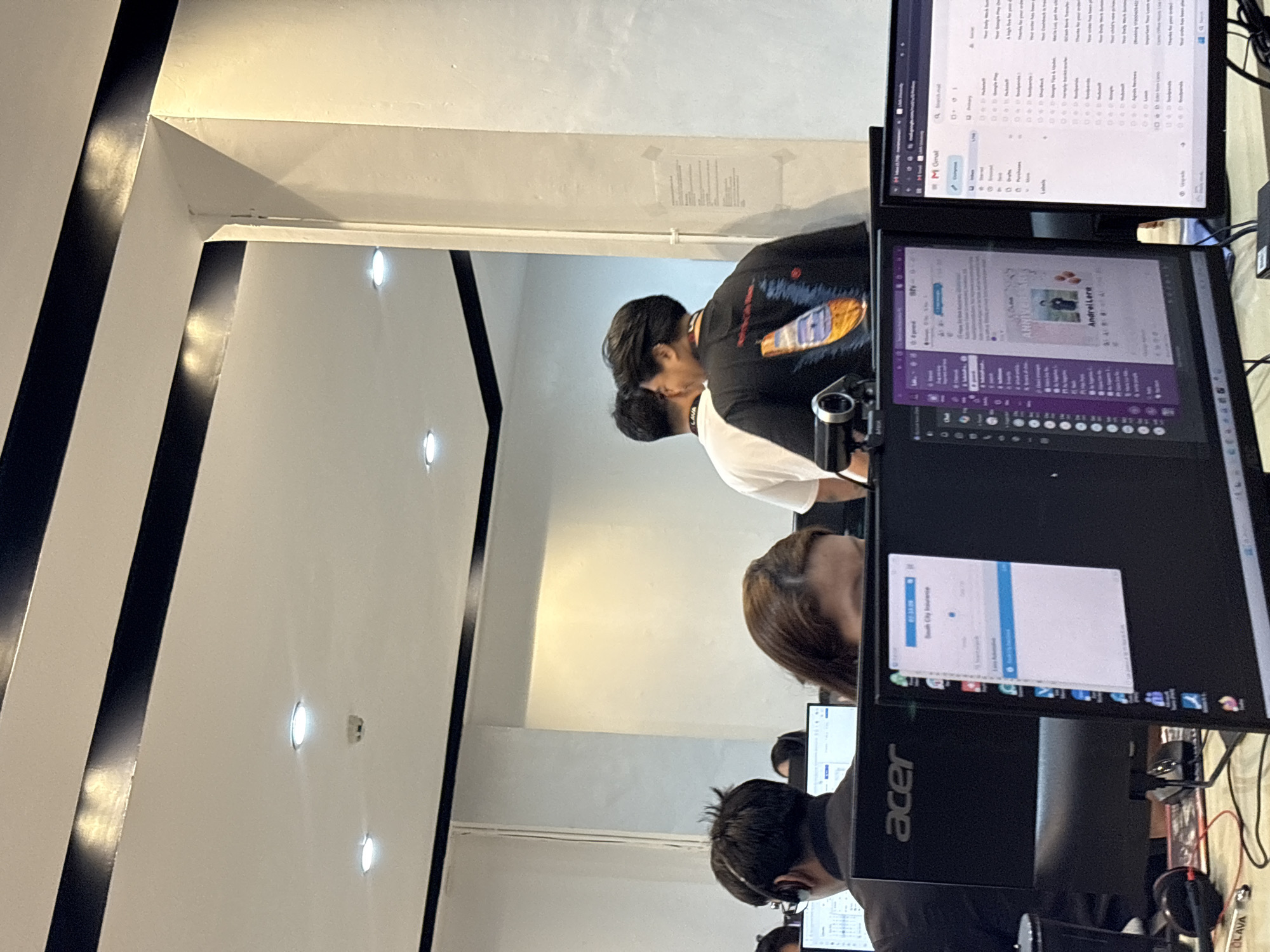 The image shows a group of people working in an office environment. In the foreground, two computer monitors are visible. The left monitor displays an "Acer" logo and a window with text, including "South City Insurance". The right monitor displays a Gmail inbox. Behind these, a woman with brown hair is visible, and behind her, two men are standing. One man is wearing a black t-shirt with a graphic print, and the other is wearing a white t-shirt. The ceiling of the room has black and white stripes, and there are recessed lights. The overall impression is of a typical office setting, possibly a call center or a tech company. There is no specific visual information that places this image in Davao City, Philippines, or any other specific location.