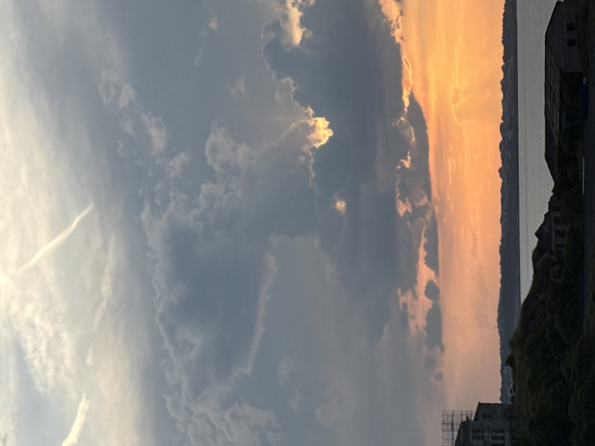 The image captures a serene coastal scene at either sunrise or sunset, with the camera held in a vertical orientation relative to the horizontal landscape. The sky dominates the left and central parts of the frame, showcasing a dramatic blend of light blue-grey clouds transitioning into vibrant orange and yellow hues on the right, indicating the low angle of the sun. Some clouds are brightly lit from behind, creating luminous edges, while others are darker and denser. A faint, wispy contrail or cirrus cloud streaks across the lighter blue sky.
Stretching horizontally across the middle-right of the image is a calm body of water, likely the Black Sea given the Sozopol, Bulgaria context, appearing in shades of grey-blue. Beyond the water, a dark, undulating landmass, possibly cliffs or hills, forms a silhouette against the brightly lit horizon.
In the immediate foreground on the right, a dark, blocky structure, resembling an old or industrial building with dark window openings, stands partially visible. Below this, dark foliage or overgrown terrain is present. Further to the left in the foreground (at the bottom of the correctly oriented image), another dark structure appears to be under construction or renovation, with scaffolding visible against the dimming light. No people are visible. The overall mood is tranquil, highlighting the natural beauty of the sky and coastline during the golden hour. No visible text is present in the image.