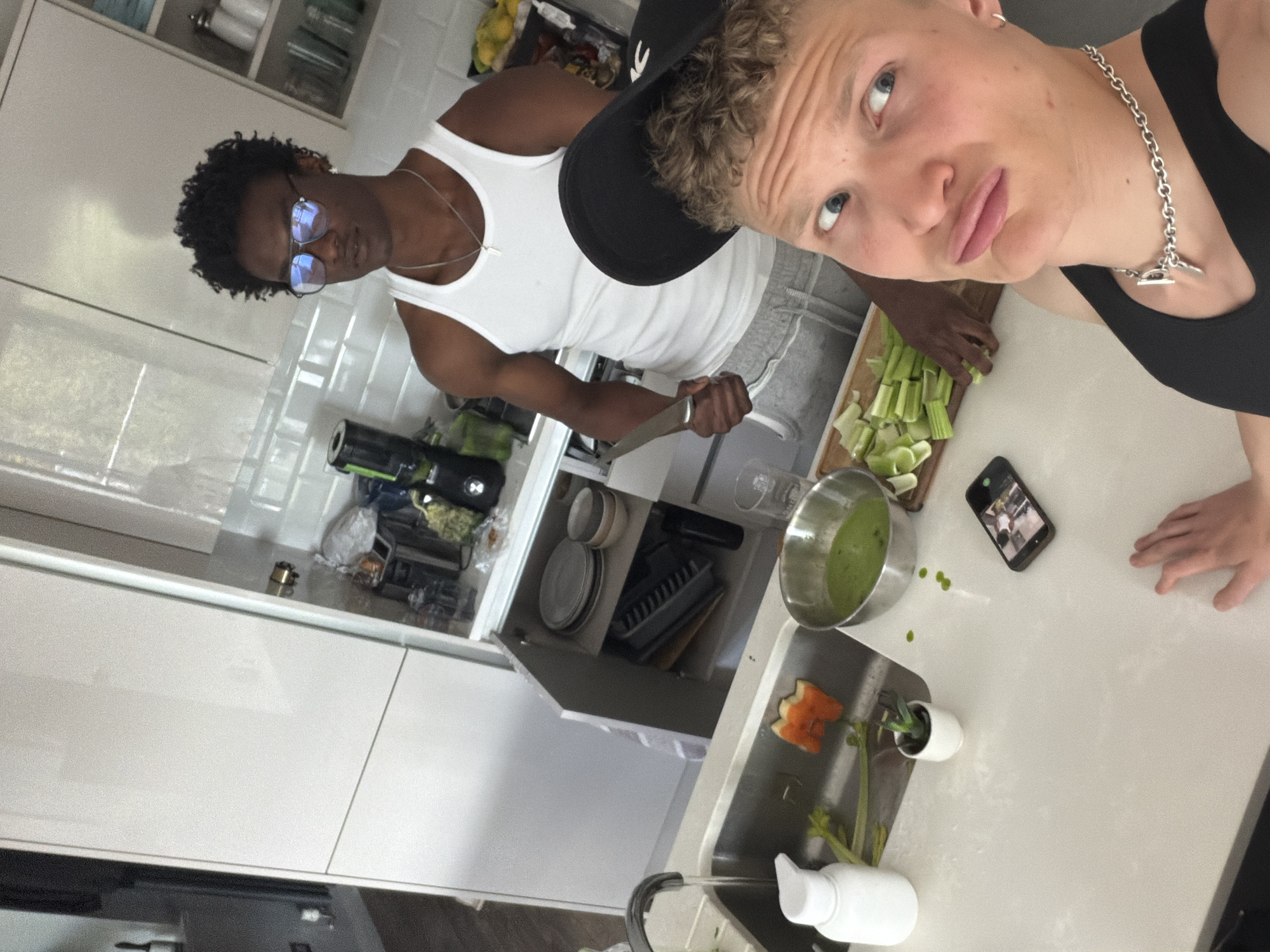 The image captures two young men in a modern kitchen, taken from a high angle and rotated 90 degrees clockwise.
In the background of the rotated image, a man with dark curly hair, wearing glasses, a white tank top, and a silver chain, holds a large knife in his right hand, looking towards the camera with a neutral expression. In the foreground, another man with short, light curly hair and blue eyes wears a black baseball cap, a black tank top, and a chunky silver chain. He looks directly at the camera with slightly pursed lips, suggesting a posed or playful expression.
The setting is a contemporary kitchen with white flat-panel cabinets and white subway tiles on the wall. On the white countertop, a wooden cutting board holds chopped celery. A stainless steel bowl contains a green liquid or puree, with small green splatters nearby. A black smartphone lies on the counter, displaying an image or video, positioned near the hand of the man in the foreground. A white soap dispenser and a small potted plant in a white pot are by a stainless steel sink, which contains what appears to be an orange peel or carved fruit, and celery stalks. An open cabinet above the counter reveals stacked plates and a dish rack.
The activity appears to be food preparation, with one man actively chopping while the other poses for the camera. The lighting indicates it is daytime. No discernible text is visible.
