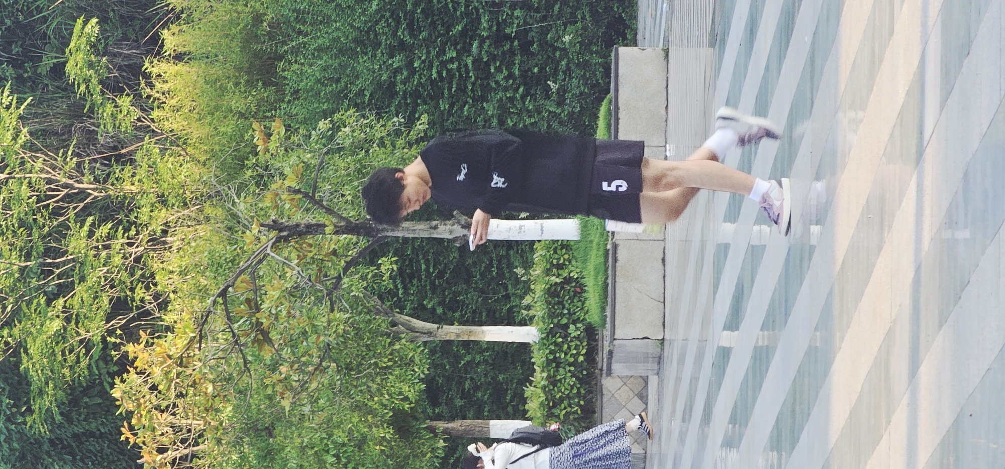 A vertical, outdoor, long-distance shot shows a person in a black shirt and black shorts with a "5" on the right leg, standing on a gray-and-white striped walkway in the city of Chongqing High-tech Zone, China. The person is looking down at a white object in their hands, possibly a phone or piece of paper. The person is standing behind a short, white-painted cement barrier. Behind the barrier, a tree with green foliage and bare branches is visible. Beyond the tree is a tall green hedge or wall of greenery. A second person, wearing a white top and long blue-and-white patterned skirt, is taking a photograph and is partially visible in the bottom left. The scene appears to be a public area.