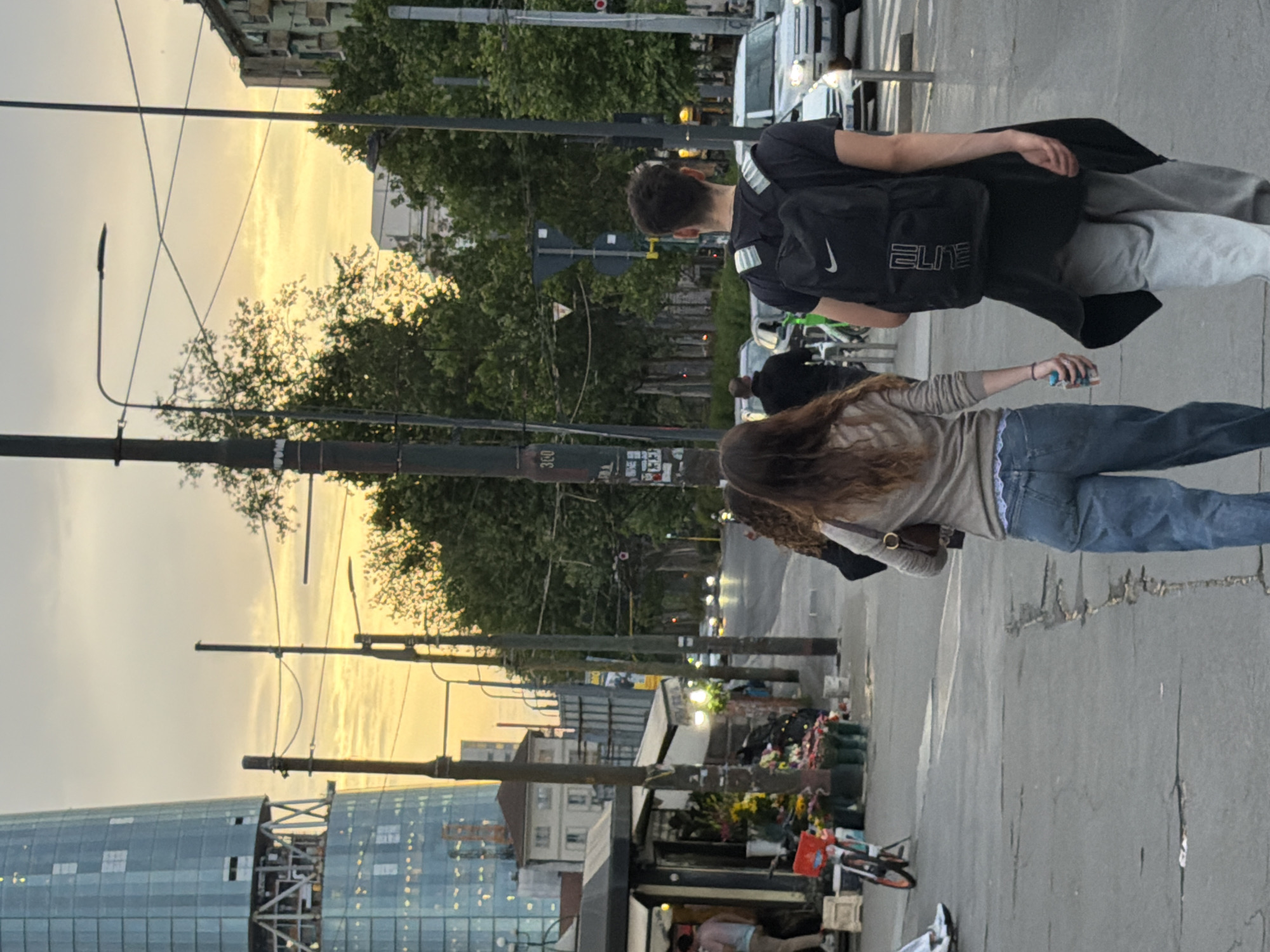 Two individuals, a male and a female, walk along a paved street. The male wears a black t-shirt with a Nike logo, a black backpack with the word "Elite" on it, grey sweatpants, and carries a black jacket over his arm. The female wears a beige sweater, blue jeans, and a brown purse on her shoulder. She holds a small, rectangular object in her hand.
The background includes a modern building with a glass facade undergoing construction, streetlights, trees, and other people. There is also a floral shop. The sky is illuminated with sunset light.
Based on the architectural style of the modern building and the street layout, the location appears to be in the city of Milan, Italy.