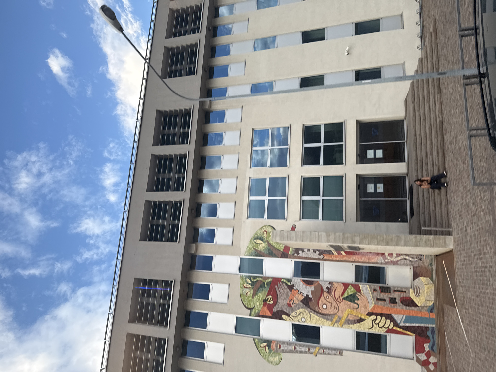The image shows a side view of a modern, multi-story building with a light-colored facade. The building has numerous windows, some of which are filled with reflective glass showing a blue sky and white clouds, while others are opaque white panels. The facade also features a large, colorful mosaic artwork depicting abstract shapes and figures, with prominent green and brown tones.
On the right side of the image, a person is visible standing on a set of stairs leading up to an entrance. The person appears to be wearing dark clothing and is looking towards the building. The stairs are made of a textured, reddish-brown material. A lamppost with a modern design extends from the top left corner of the frame, arching over the building.
The scene is set outdoors, under a bright blue sky with scattered white clouds, suggesting a daytime setting with fair weather. The orientation of the image is such that the sky is on the left and the building occupies the right portion. The architectural style and the mosaic art suggest an urban or institutional setting. No visible text is present in the image.