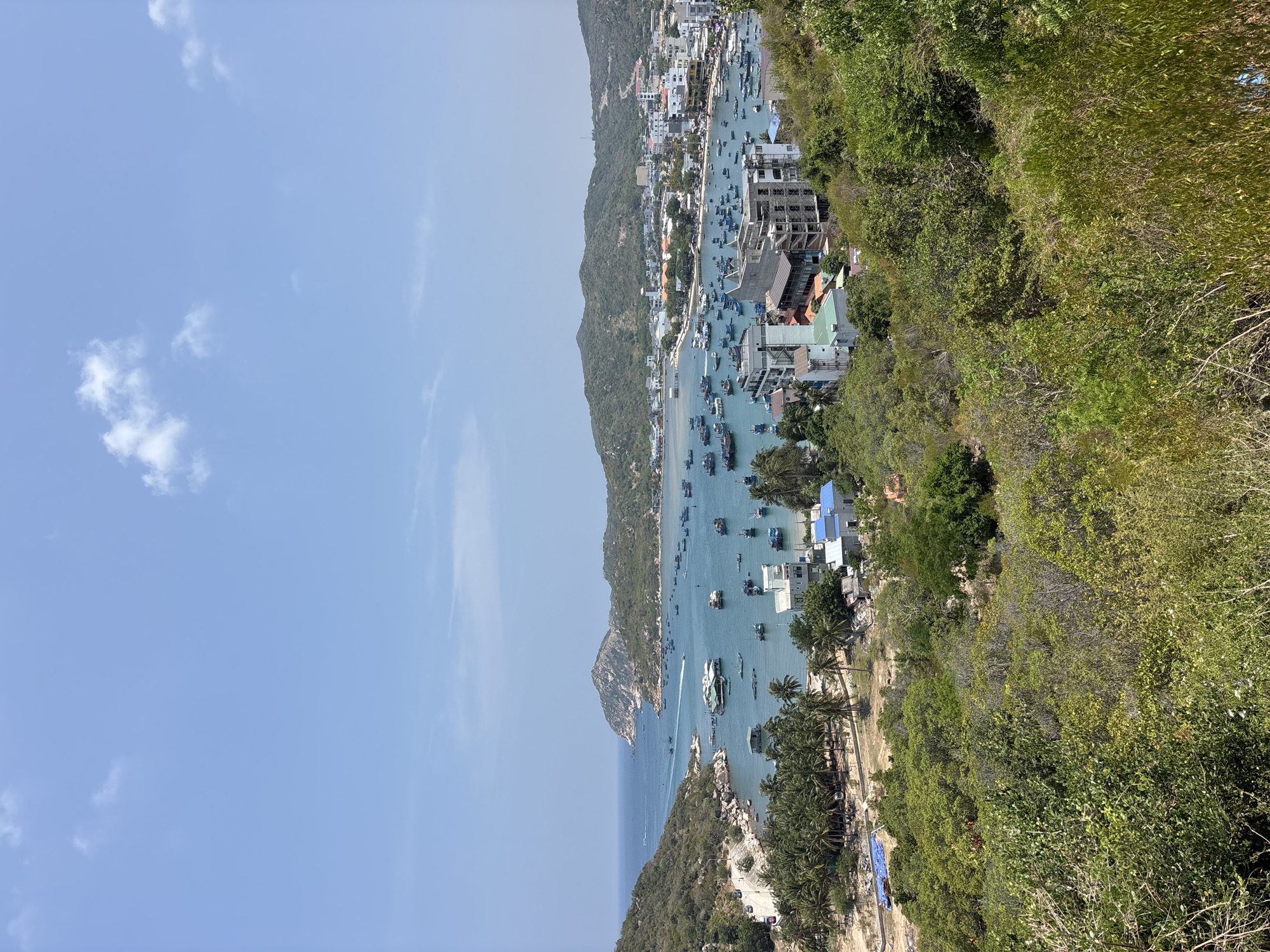 The image shows a coastal scene in Vĩnh Hải, Vietnam, likely taken from an elevated viewpoint. The sky is a clear blue with sparse cloud cover. A harbor or bay is visible, filled with numerous boats of varying sizes. Along the shoreline, buildings and structures appear densely packed, indicative of a town or settlement. The surrounding landscape consists of lush, green vegetation, and rocky outcrops. The overall scene depicts a bustling coastal community nestled within a natural environment.
