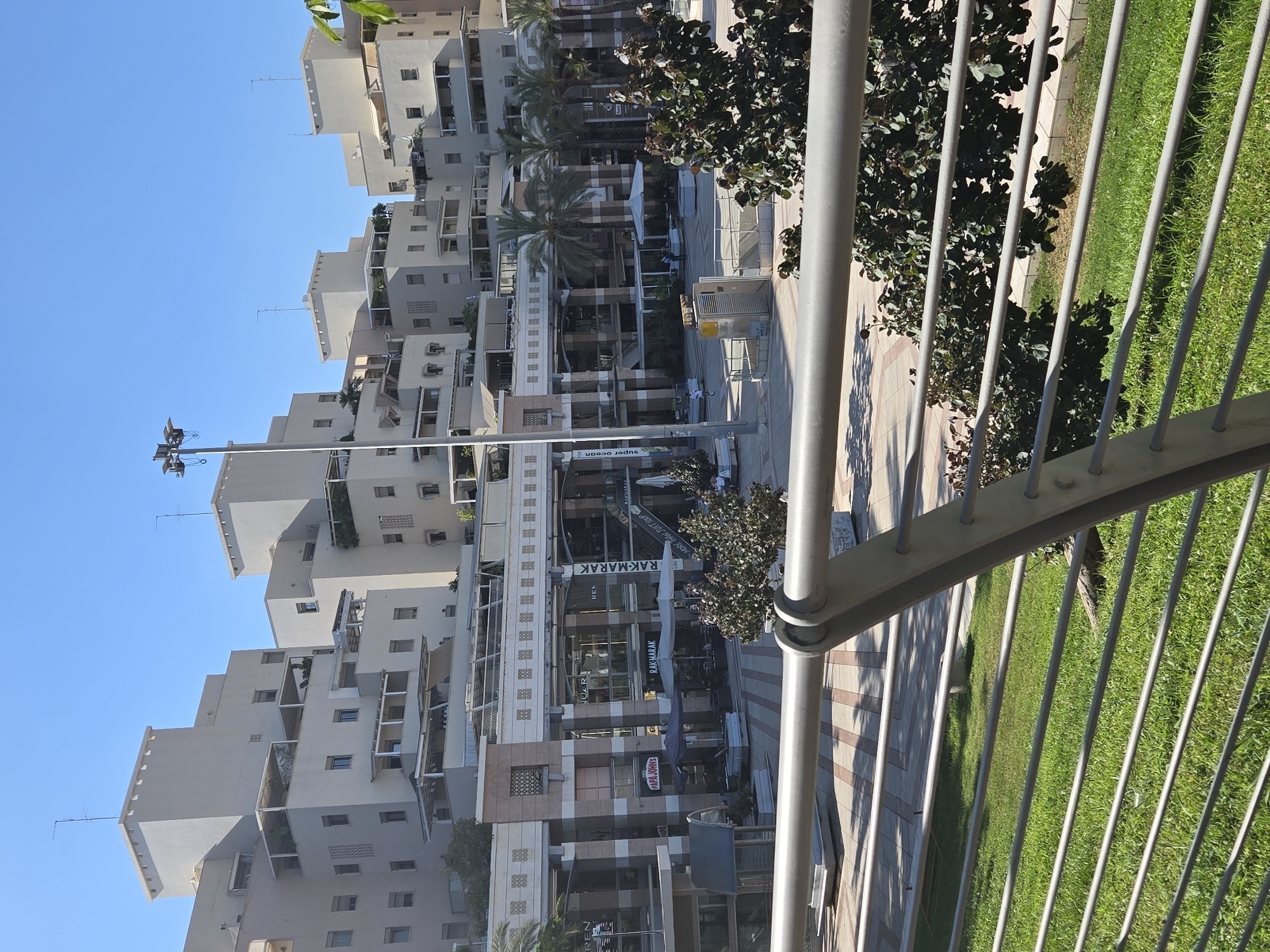 This media file depicts a bright, sunny day in a commercial area, likely Kfar Saba, Israel. The scene is dominated by modern architecture, featuring cream-colored buildings with multiple levels, balconies, and balconies with potted plants. A prominent metal railing runs across the foreground, suggesting the viewpoint is from an elevated position, such as a balcony or walkway.
Below the buildings, a pedestrian walkway is visible, paved with light-colored tiles. Shops are situated on the ground floor of the buildings, with visible signage including "PAPA JOHNS", "RAK-MARAK", and "super ocean." Palm trees add a tropical element to the urban landscape. The sky is a clear, vibrant blue, indicating fair weather.
No specific activity or event is clearly discernible, though the presence of shops and walkways suggests a public space. A few people can be faintly seen walking on the pavement. The overall impression is one of a well-maintained, modern urban environment bathed in the warm light of day.