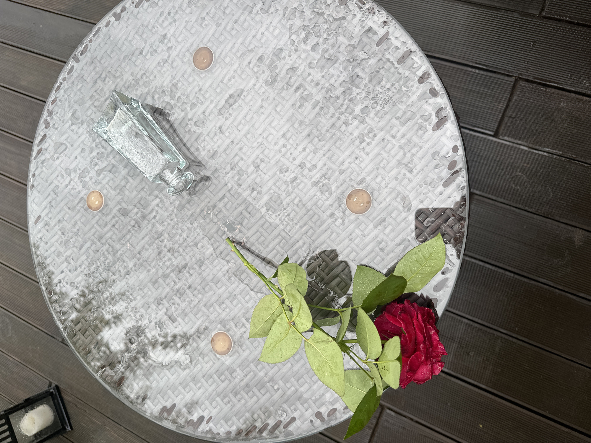 A round glass-topped table with a wicker-like pattern is situated outdoors on a wooden deck. The table surface is wet with scattered water droplets and puddles, suggesting recent rain. On the table, there is a single red rose with green leaves, a small rectangular glass vase, and four small tealight candles in holders. In the upper left corner of the image, a dark lantern with a white candle inside is visible. The setting appears to be a peaceful, possibly garden or patio, area. The location is identified as Staritsa, Russia.