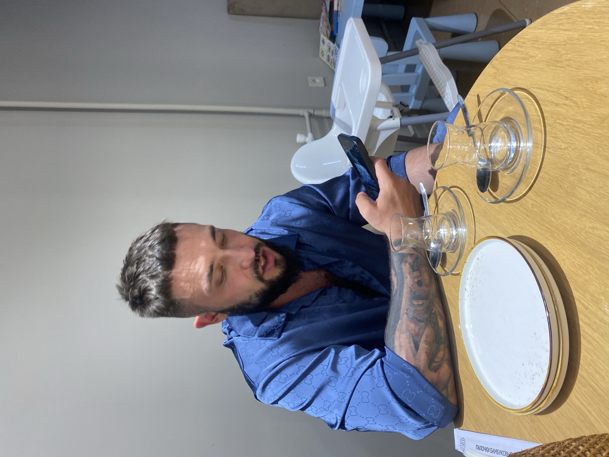 The image shows a man sitting at a wooden table in what appears to be a cafe or restaurant. He is wearing a blue patterned button-up shirt and looking at his phone. On the table in front of him are two empty tea glasses with saucers and spoons, and a white plate. In the background, a white high chair for a baby is visible. The overall environment appears to be indoors, with plain grey walls. The location is stated to be Makhachkala, Russia.
