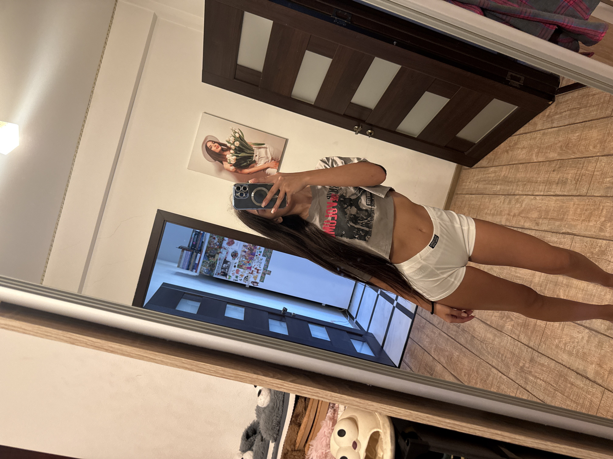 A young woman is taking a mirror selfie in a room with wooden flooring and dark brown doors. She is wearing a gray t-shirt and white shorts. The t-shirt has a graphic design on it, and the shorts appear to be athletic wear. In the background, a framed picture of a woman with a bouquet of white tulips hangs on the wall. Another reflection in the mirror shows a doorway with shelves filled with books and other items. The overall impression is of a casual, indoor setting. The image appears to have been taken during the day, as indicated by the lighting. The location is identified as Chișinău, Moldova.