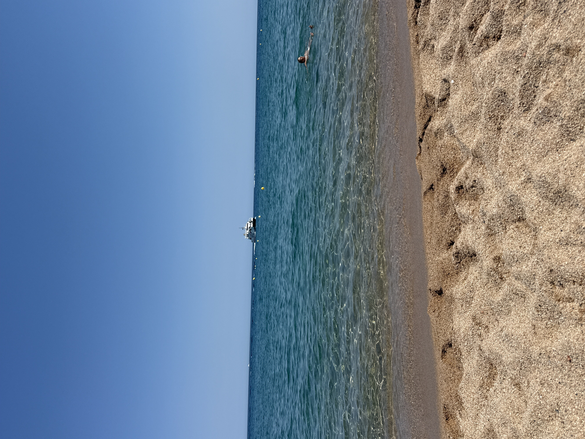 The media file depicts a serene beach scene in Blanes, Spain. The image is oriented vertically, with the clear blue sky occupying the left side. The ocean, a vibrant turquoise, stretches across the middle, its surface rippling gently under the sunlight. A person is visible swimming in the water, their body partially submerged, suggesting a leisurely activity.
In the distance, a white boat with several decks is anchored. Several yellow buoys are scattered across the water, possibly marking a swimming area or navigational channel. The right side of the image showcases the sandy beach, with footprints visible in the coarse, light brown sand, indicating recent activity. The sun appears to be high in the sky, casting a bright light on the scene, suggesting it is likely midday. The weather seems pleasant, with clear skies and calm waters, ideal for a day at the beach. The overall mood is one of tranquility and relaxation, capturing the essence of a summer day by the sea. No visible text is present in the image.