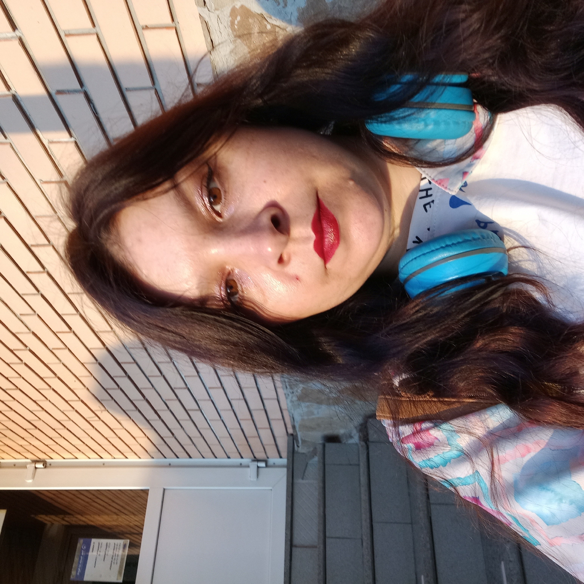 A medium shot selfie captures a woman with long, dark brown hair and brown eyes, looking directly at the camera. She is wearing red lipstick and blue headphones around her neck. Her clothing features a colorful, abstract pattern of pink, blue, and white. The background consists of a brick wall, a white door with a sign on it, and a set of grey stairs. The lighting suggests it is late afternoon, with sunlight casting shadows on the brick wall. The scene appears to be outdoors, possibly in front of a building or entrance.