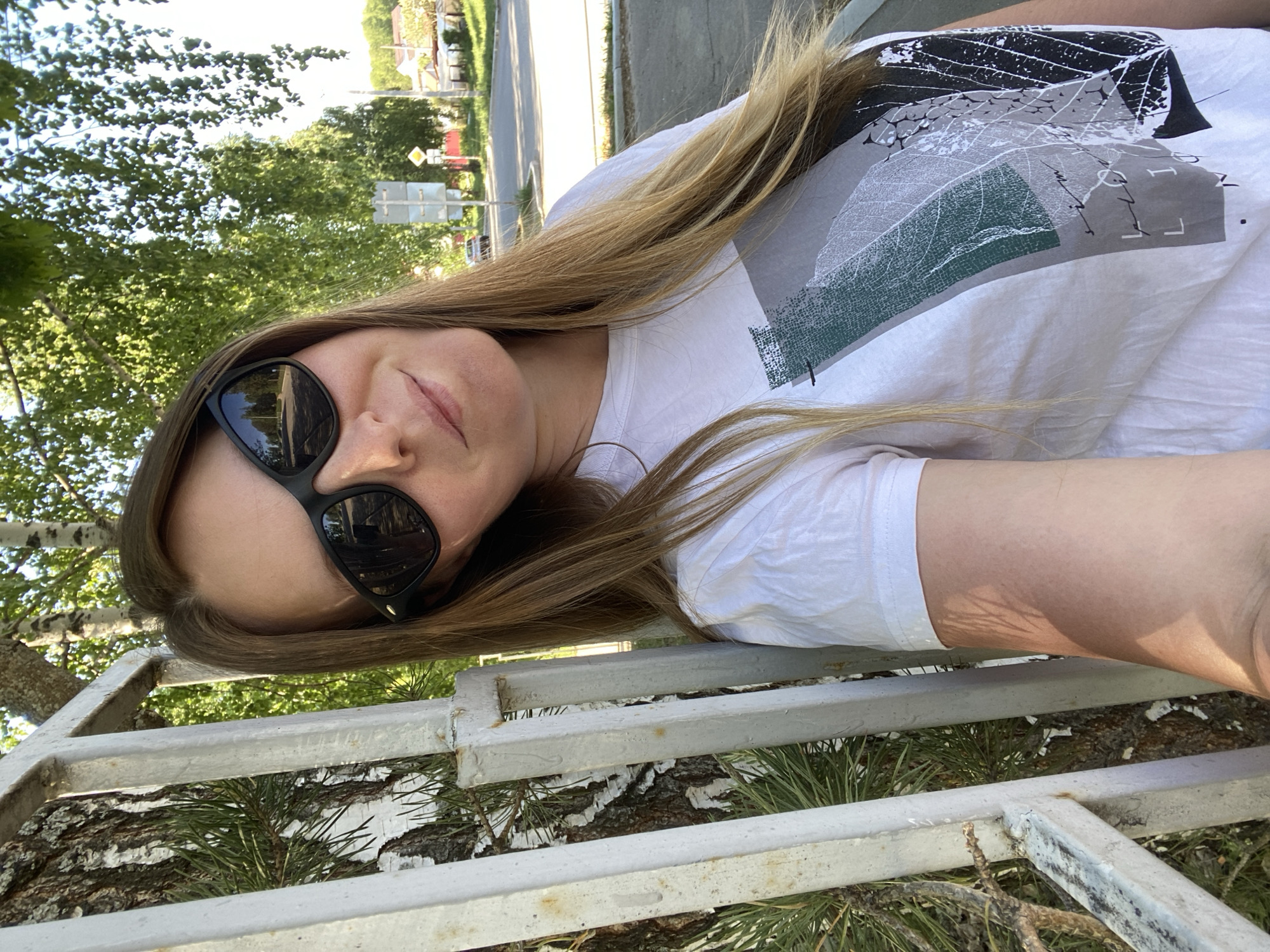 A woman with long blonde hair and sunglasses is posing for a selfie. She is wearing a white t-shirt with a leaf design. She is leaning against a white metal fence. Behind the fence is a tree with white bark. In the background, there is a road with a yellow diamond traffic sign and buildings. The location is Noviy, Russia.