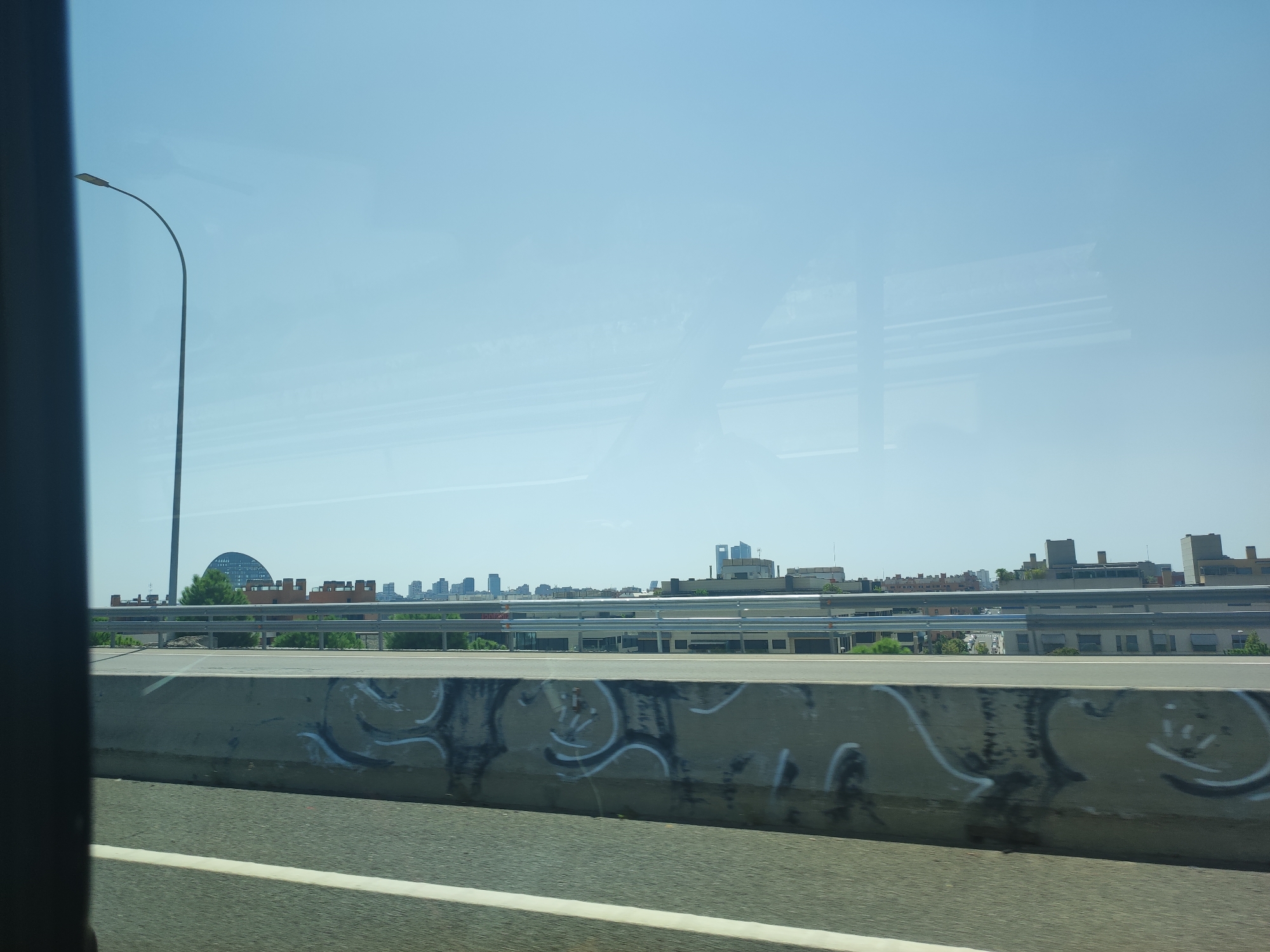 The image shows a view from a highway in Madrid, Spain. In the foreground, a concrete barrier with graffiti is visible, along with a road with white lane markings. Beyond the barrier, a metal guardrail runs parallel to the road. In the middle ground, a cityscape with various buildings and trees is seen. A distinctive arched building with a dark facade is prominent on the left side of the city skyline. A tall street lamp is positioned on the left side of the image, with its curved arm extending over the road. The sky is clear and bright blue. The image appears to be taken during daylight hours.