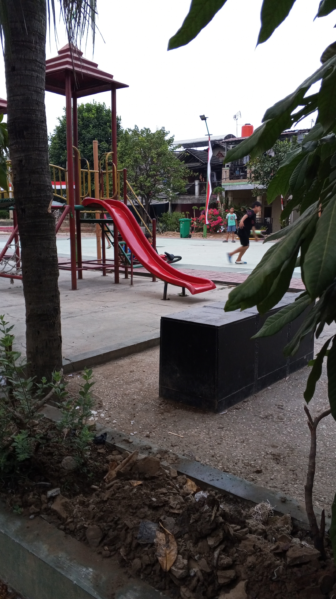 An exterior, eye-level, medium shot captures a playground in Bekasi, Indonesia. A red slide dominates the foreground, attached to a larger red playground structure with a peaked roof and yellow railings. To the left, the textured trunk of a palm tree stands tall, partially obscuring the playground. In the middle ground, two children are running. One child, wearing a blue shirt and dark shorts, is in mid-stride, while another child in a black shirt and shorts runs in the opposite direction. Behind them, several buildings with peaked roofs are visible, adorned with red and white flags. Green foliage surrounds the area, and a trash can is situated to the right of the slide. A dark, rectangular bench sits in the foreground, partially covered by leafy branches. The ground is paved with concrete and gravel.