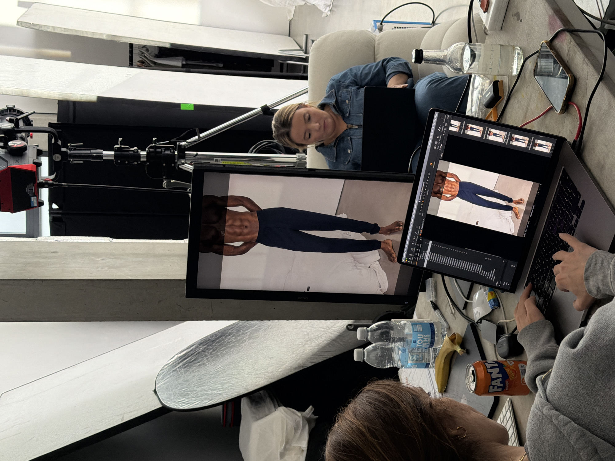 The image depicts a behind-the-scenes view of a photo shoot, likely in a studio setting. Two women are visible. One woman, wearing a denim jacket, is partially visible, reclining on a white sofa. A second woman, with brown hair, is hunched over a laptop, typing. Two monitors are positioned in front of them, displaying images of a male model wearing blue leggings. A professional camera setup is visible to the left. The surrounding environment includes photography equipment such as reflectors and lighting. Items on the workspace include water bottles, a banana, and a can of Fanta soda. There is no identifiable information linking the scene to London, United Kingdom.