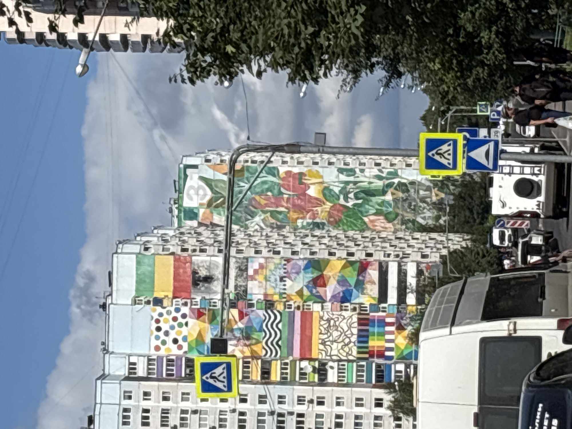 The image depicts a street scene in Odintsovo, Russia, dominated by a brightly painted residential building. The building's facade is adorned with a vibrant mural featuring abstract geometric shapes, polka dots, wavy lines, and floral motifs in a multitude of colors, including blue, green, yellow, red, purple, and orange. The building itself appears to be a multi-story apartment complex, with visible windows and balconies.
In the foreground, a white van is parked on the right side of the road, facing away from the camera. To the left of the van, the rear of a dark-colored car is partially visible. The street also shows traffic signs, including a pedestrian crossing sign (a blue square with a yellow border and a white pedestrian silhouette) and a speed limit sign (indicating 20). Further down the road, other vehicles, including a white bus and a white utility vehicle, are visible.
Several people are walking on the sidewalk on the right side of the street, some carrying bags. Green trees line the right side of the road, partially obscuring another tall building in the background, which appears to be another apartment block with a more conventional facade. The sky is a bright blue with scattered clouds. The overall impression is a well-populated urban environment with a visually striking architectural enhancement.