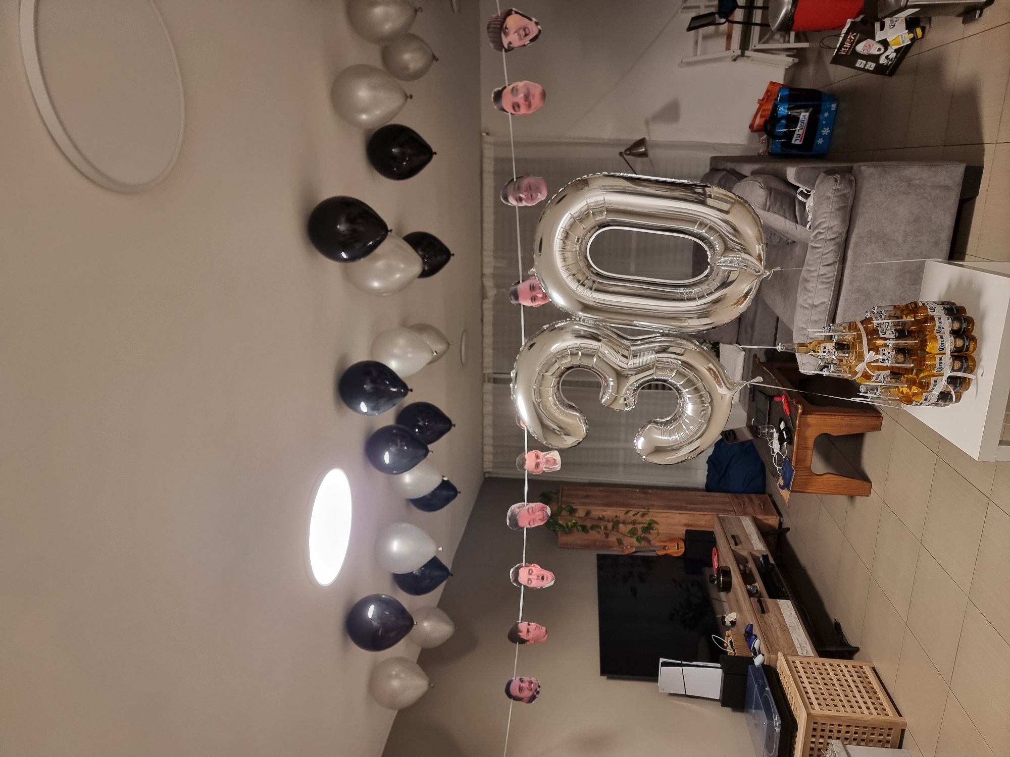 The image depicts a domestic indoor setting, likely a living room, decorated for a birthday celebration. A large number "30" formed by silver foil balloons dominates the foreground. Above, a string of balloons in black and silver hangs from the ceiling, interspersed with a garland of printed faces. The room contains furniture such as a television stand with a TV, a sound system, and a gaming console, a gray sofa, and a wooden coffee table with laptops and drinks. In the background, there are shopping bags and chairs, suggesting a home environment. The overall scene is set in Ramat Gan, Israel, indicated by the Hebrew text on one of the shopping bags. No people are visible in the image.