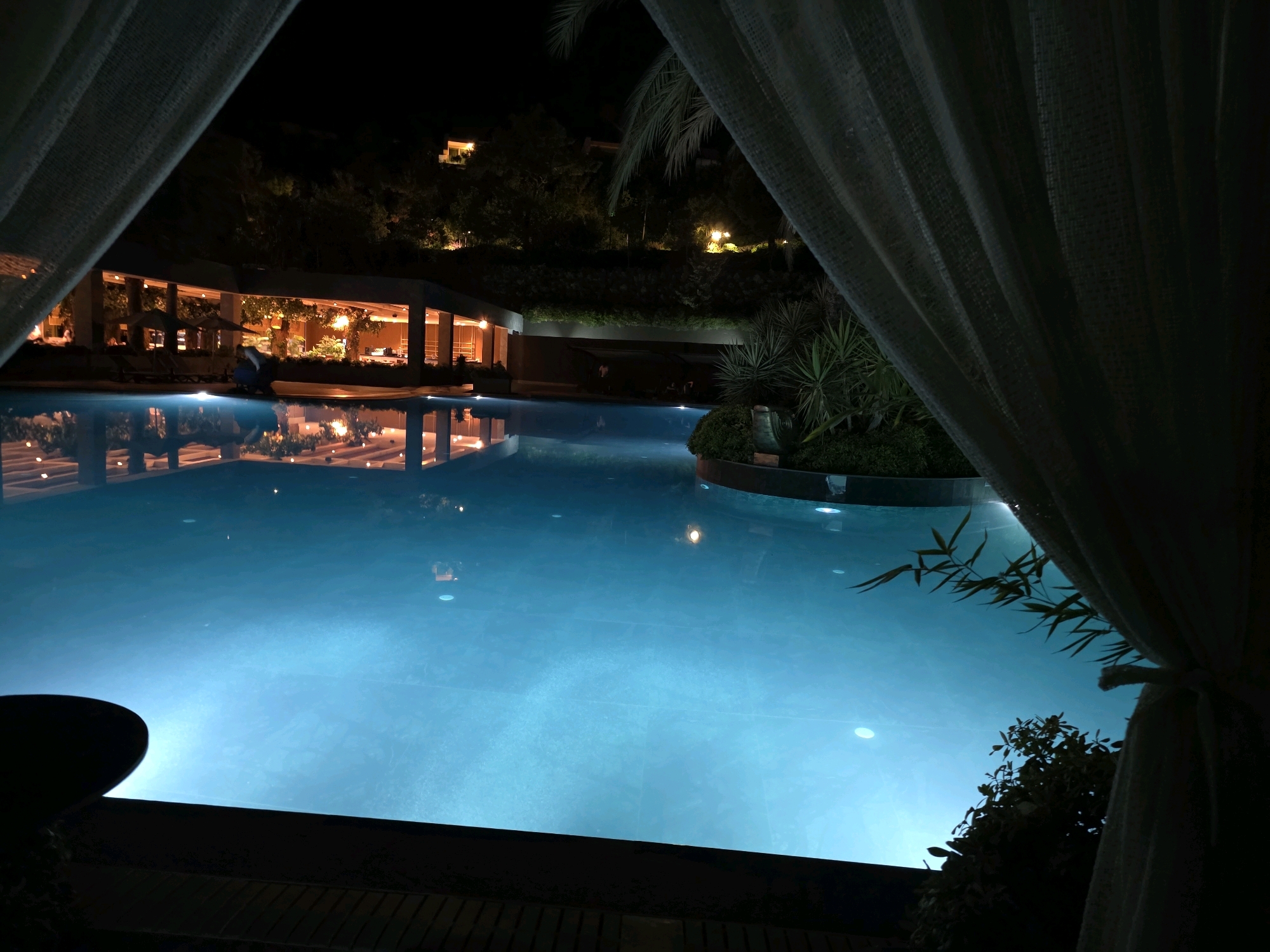 A nighttime view of a well-lit swimming pool and an outdoor restaurant area is framed by sheer white curtains, suggesting a resort or hotel setting in Bodrum, Turkey. The pool's bright turquoise water is illuminated from within, casting reflections on the underside of the draped fabric. Beyond the pool, an open-air dining area with illuminated tables and chairs is visible, backed by lush greenery and a hillside dotted with lights. The overall atmosphere is tranquil and inviting, characteristic of a luxury vacation destination.