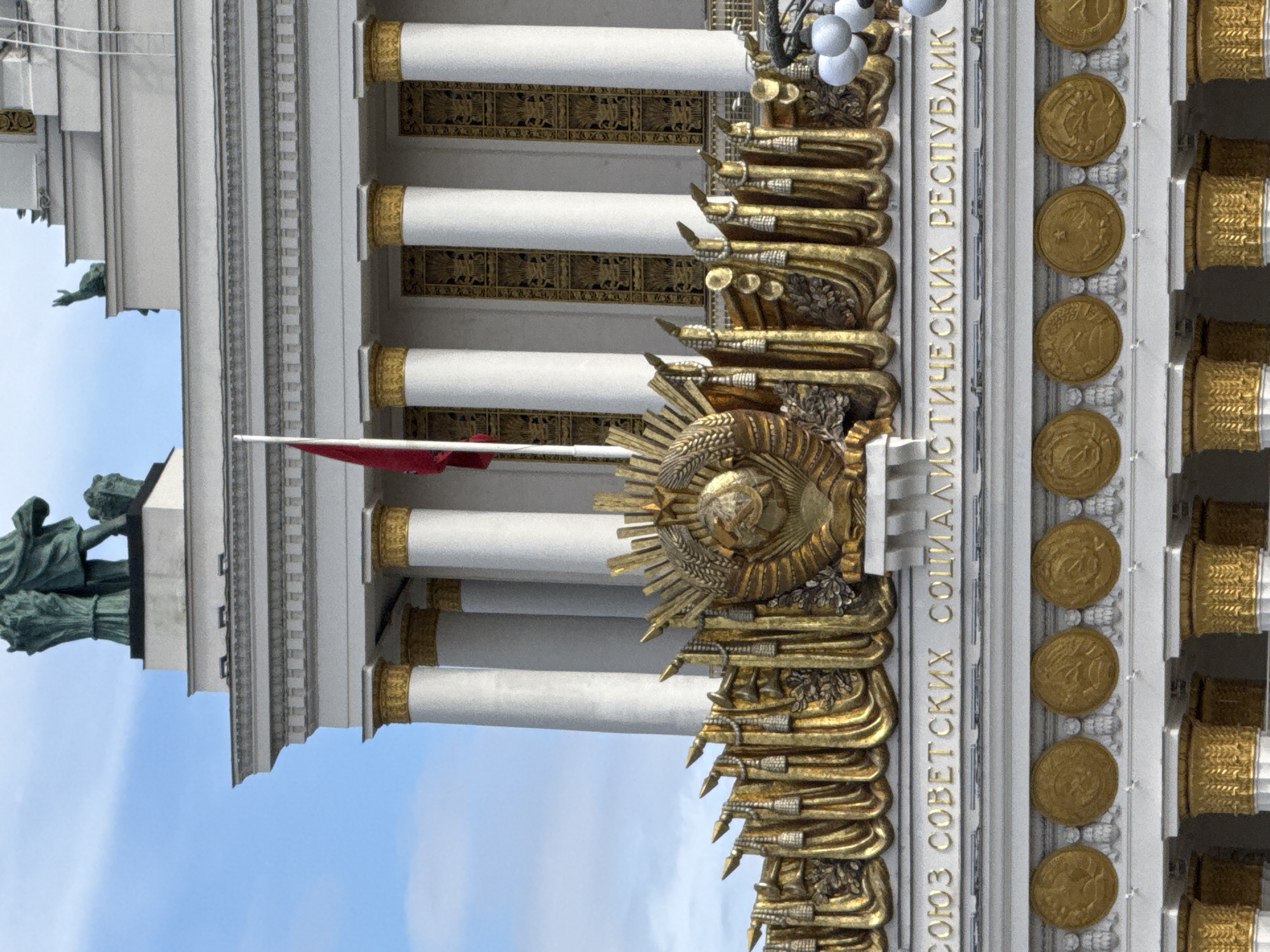 The image shows a detail of a building's facade in Moscow, Russia, specifically the VDNKh (Exhibition of Achievements of National Economy) complex. The focus is on the upper portion of the building, featuring classical architectural elements. Dominating the scene are large, white, fluted columns topped with ornate gold capitals. Between the columns, the wall is decorated with intricate gold friezes and panels featuring Soviet-era symbols.
A prominent gold emblem, the hammer and sickle with a star and wheat stalks, is centrally placed within a circular frame, flanked by more gold decorative elements resembling garlands and military standards. Below this emblem, in gold lettering, is text that reads "СОЮЗ СОВЕТСКИХ СОЦИАЛИСТИЧЕСКИХ РЕСПУБЛИК" (Union of Soviet Socialist Republics). Beneath the inscription, a series of circular medallions, also in gold, are mounted on the facade, each appearing to depict a Soviet emblem or scene.
A red flag flies from a flagpole on the left side of the image, partially obscuring some of the architectural details. The sky above is a clear blue with some wispy clouds. The overall impression is one of grandeur and Soviet symbolism, characteristic of the architecture found at VDNKh.