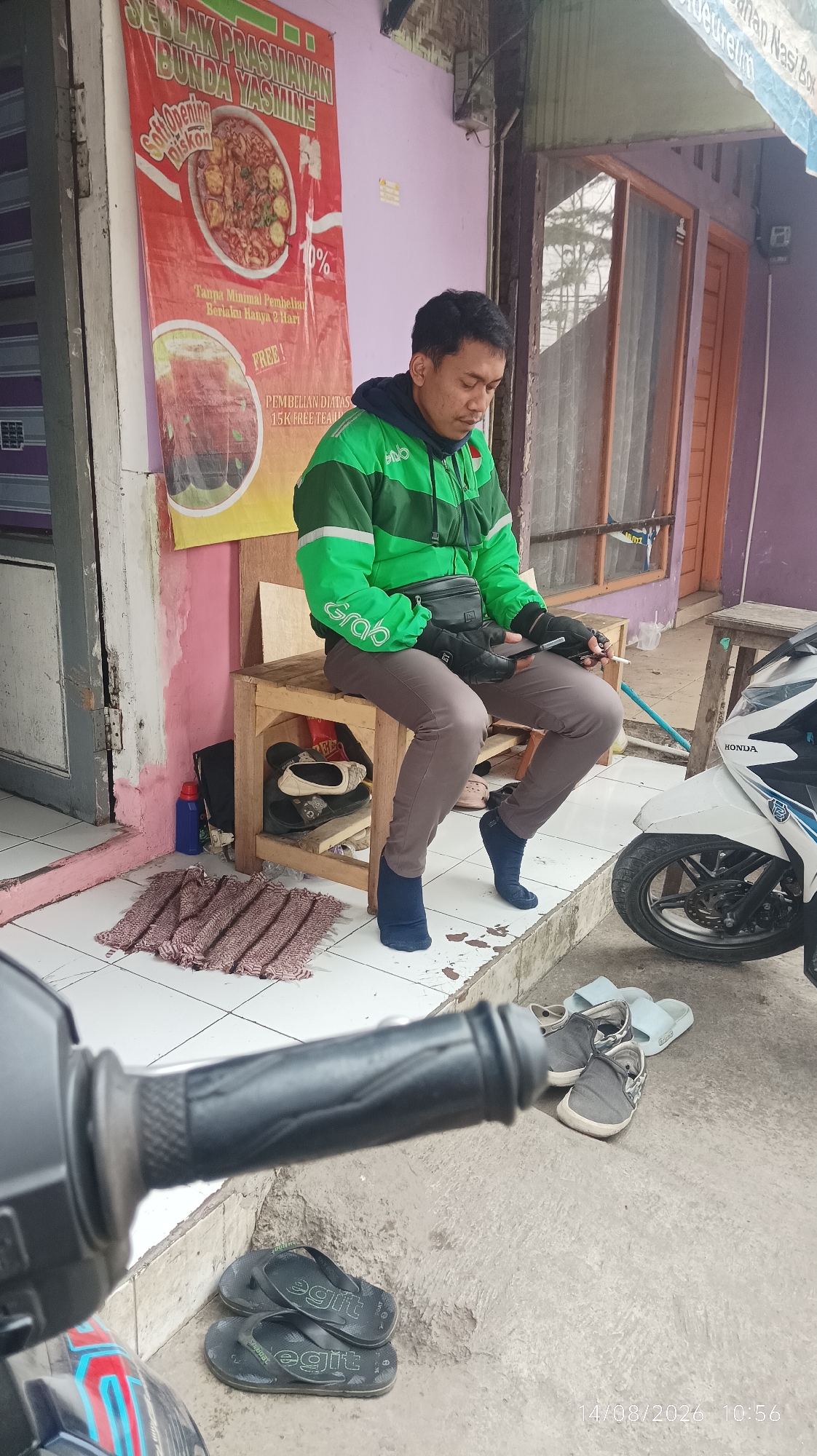 A man wearing a green Grab jacket and grey pants is sitting on a wooden chair. He is holding a phone and a cigarette. In front of him is a motorcycle, and there are several pairs of shoes and sandals on the ground. Behind him is a banner advertising "Seblak Prasmanan Bunda Yasmine" with a discount. The scene appears to be on a street in Tasikmalaya, Indonesia.