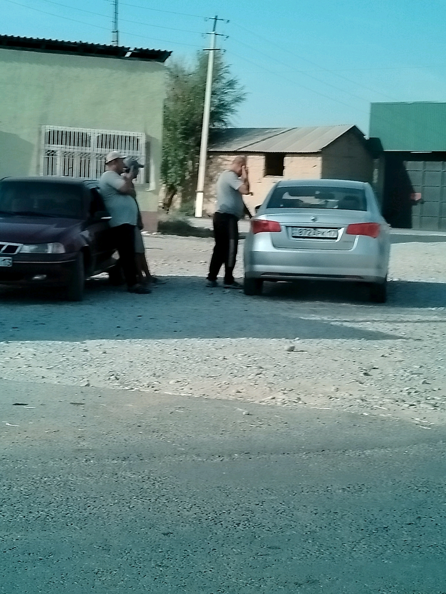 The image depicts an outdoor scene in the city of Kölkent, Kazakhstan. In the foreground, a dirt and gravel road is visible. Two men are standing next to a maroon car on the left side of the frame, and a silver sedan is parked in the middle ground to the right. Behind the vehicles, a light green building with barred windows is on the left, and to the right, there are several smaller structures with corrugated metal roofs. Power lines and utility poles are present in the sky. One man, wearing a light-colored t-shirt and cap, is leaning against the maroon car and appears to be holding something to his ear, possibly a phone. The second man, in a grey t-shirt and black pants, is standing near the trunk of the maroon car. The silver sedan has a license plate with Cyrillic characters that reads "B72EX 17". The overall impression is that of a quiet, possibly rural or industrial area.