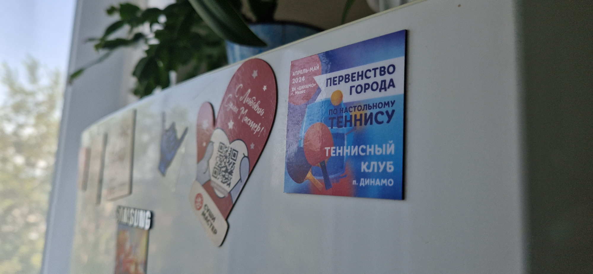 The image shows a close-up of a white refrigerator door with several magnets and stickers. On the left, there is a heart-shaped magnet with a QR code and text in Russian that appears to be related to a sushi restaurant. To the right of this, there is a sticker advertising a city championship in table tennis, with the dates April-May 2024, and mentioning DK "Dynamo" in Miass. The sticker also features images of table tennis rackets and balls, and the text "TENNIS CLUB". The background shows a plant and a window with sunlight streaming in, creating a shadow on the refrigerator door. There are no people or animals visible in the image.