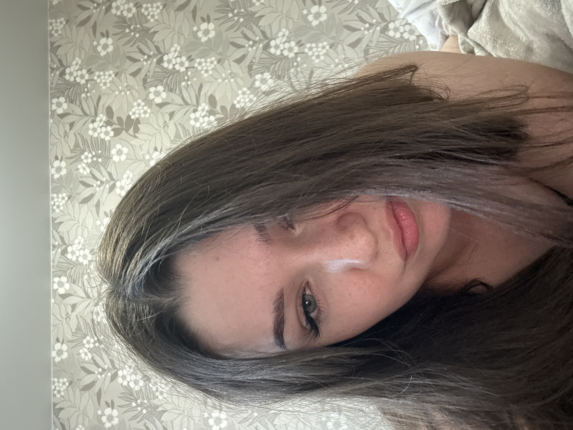 The media file shows a close-up, head-and-shoulders shot of a young woman lying down, with her head tilted to the side and her dark hair obscuring part of her face. She appears to be indoors, with patterned wallpaper featuring white flowers on a grey background visible behind her. Her skin has a slight glow, and her eyes are open and looking towards the viewer, with long eyelashes. Her lips are parted, revealing a hint of teeth. The lighting suggests it could be daytime, possibly in the morning or afternoon, given the soft, natural-looking illumination. The overall mood is intimate and relaxed, with the woman's pose and direct gaze creating a personal connection. There is no visible text in the image.