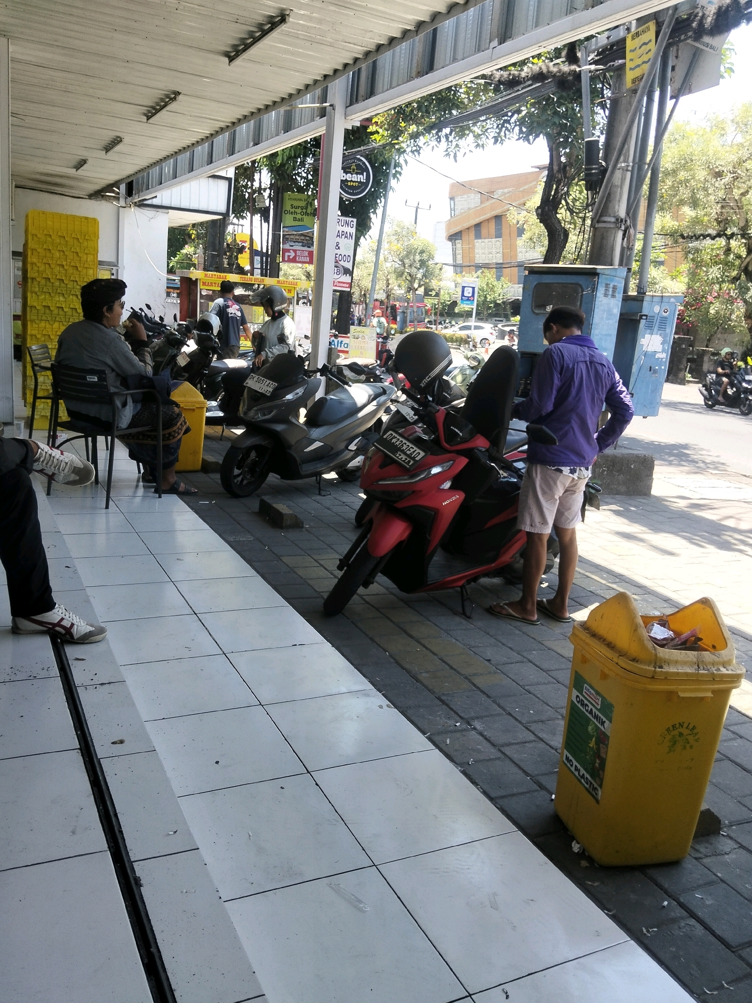 The image shows a street scene in Kuta, Indonesia, likely in front of a storefront or restaurant. In the foreground, a person is seated on a chair, facing left and eating. To their right, a man in a purple shirt and light-colored shorts stands near a blue electrical box, looking down. A red and black scooter is parked in front of him, with a black scooter parked behind it. To the right of the scooters is a yellow trash bin with "ORGANIK NO PLASTIC" labels. The ground is paved with white tiles in the foreground and dark gray pavers further back. In the background, there are buildings, trees, street signs, and other parked motorcycles. The overall impression is a bustling street with typical urban elements.