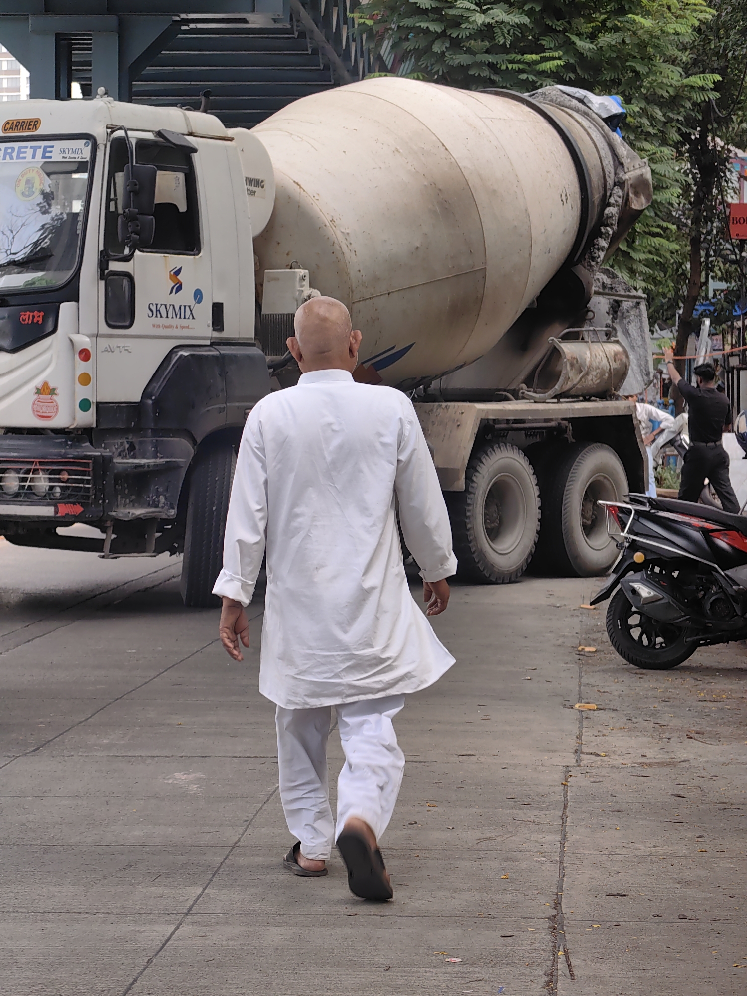 A bald man wearing a white kurta and white pants walks away from the camera on a paved street in Mumbai, India. He is wearing dark sandals. Beside him and to his left is a white cement mixer truck with "SKYMIX" written on its side. The truck has a large rotating drum. To the right of the truck and partially in front of it, a black scooter is parked. In the background, there are trees and an elevated walkway or bridge with blue railings. A man in a black uniform is visible in the background, standing near the truck and looking towards the left. The overall scene appears to be an urban environment with construction activity.