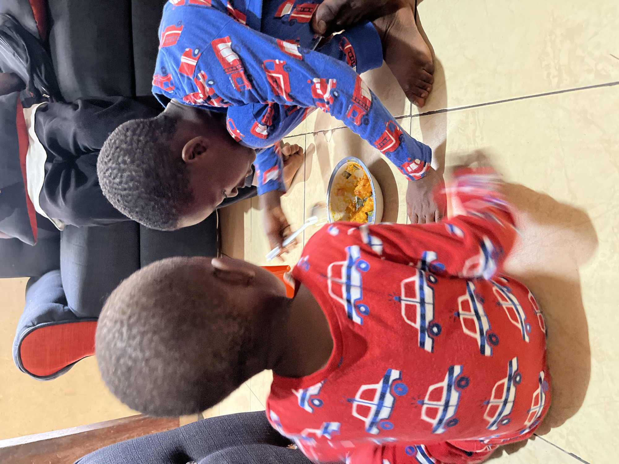Two young children in pajamas sit on the floor and a couch eating from a bowl. One child is wearing blue pajamas with a red firetruck pattern, and the other is wearing red pajamas with a blue car pattern. The location appears to be indoors on a tiled floor, with a couch in the background. The specific location is Badagry, Nigeria.