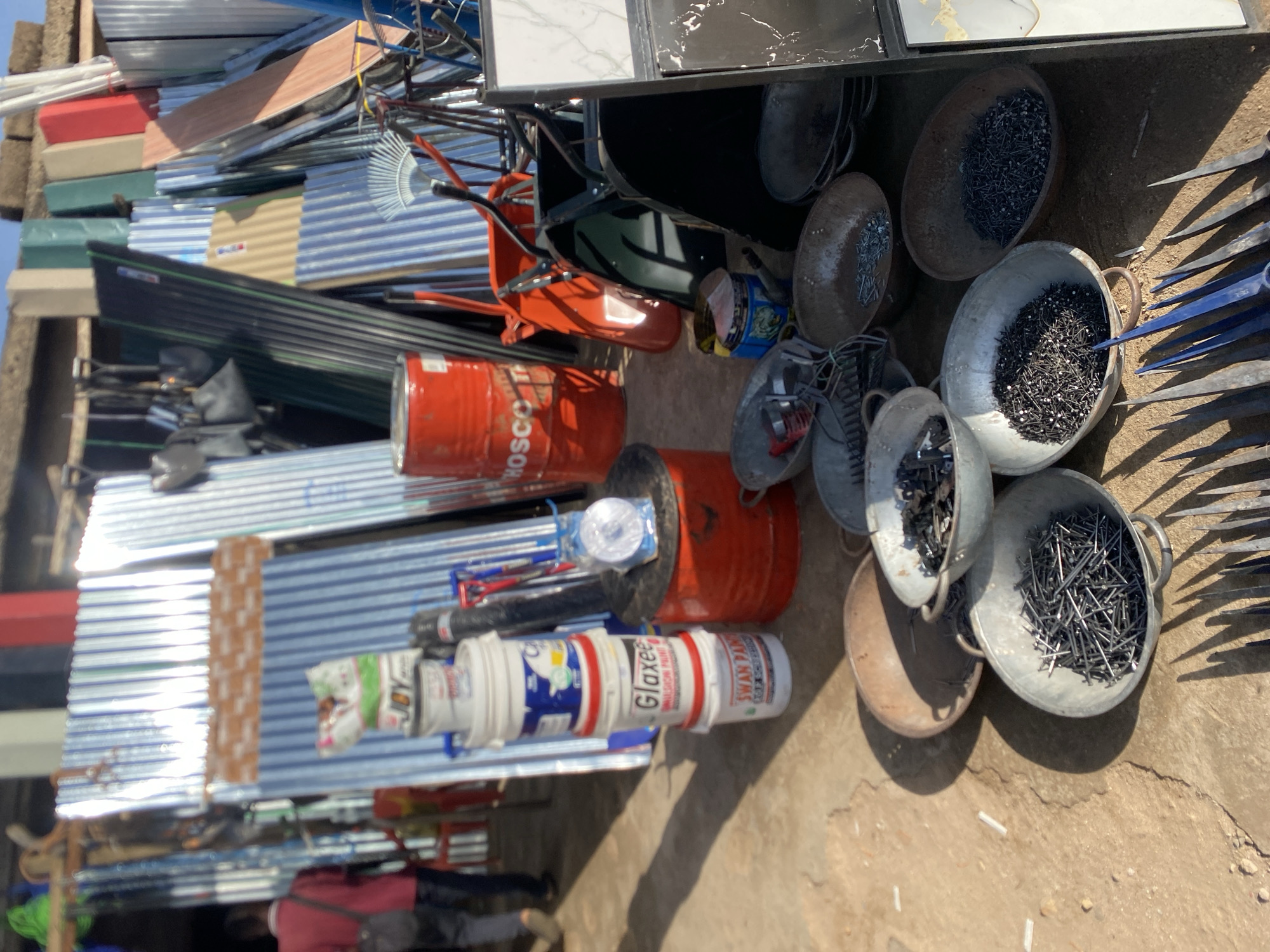 The image shows an outdoor market scene in Bukuru, Nigeria. A variety of construction materials are visible, including stacks of corrugated metal sheets, paint cans (brands such as "Glaxee" and "Swan Paints" are discernable), and red metal barrels, one labeled "Hosco." Several metal bowls containing nails are on the ground, along with some blue metal tools. The image seems to have been taken during the daytime. The focus is on the market inventory, with a glimpse of people's legs in the background.