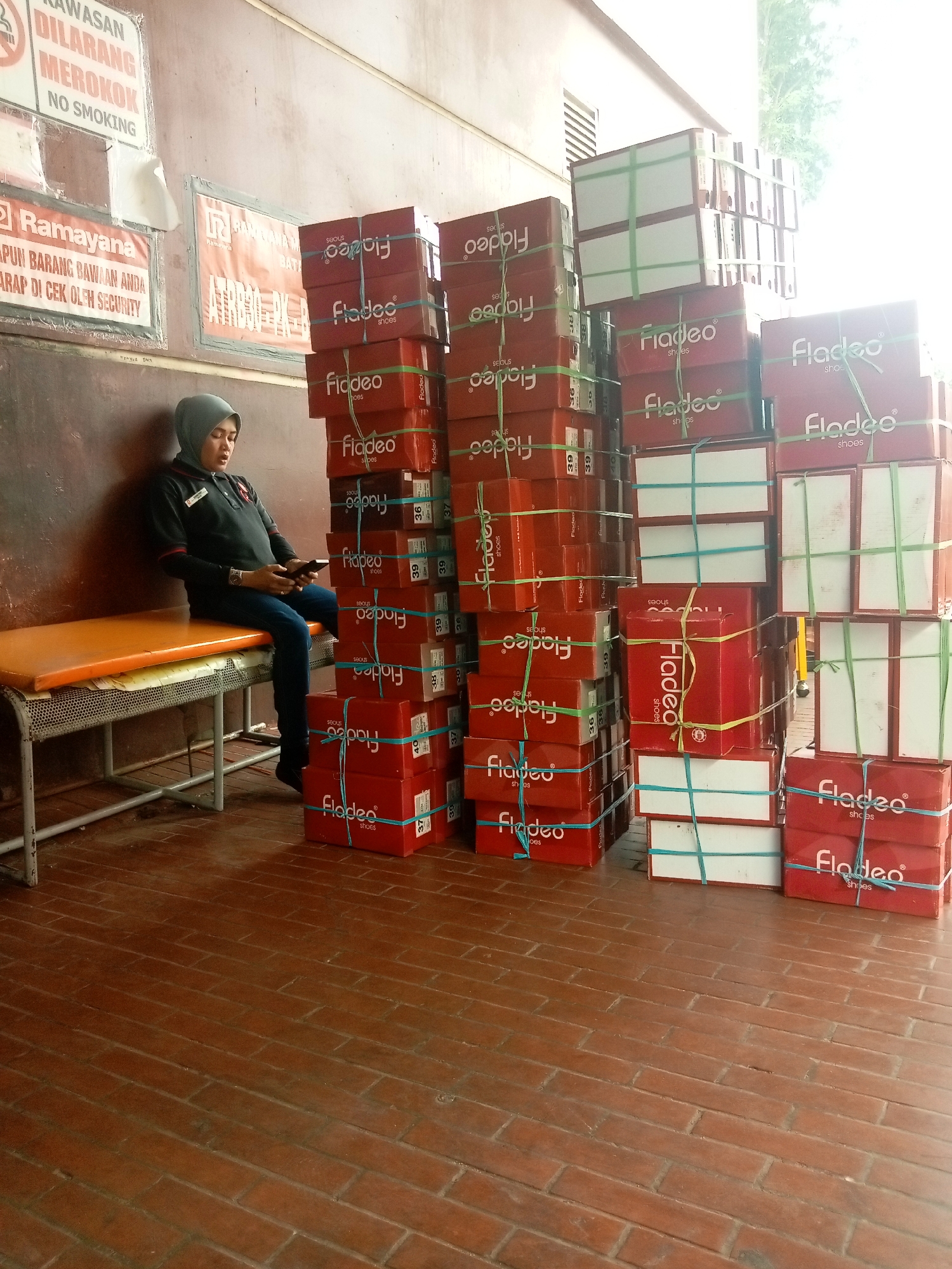 A woman wearing a gray hijab and a black shirt with a red collar is sitting on an orange bench, looking at her phone. The bench is in front of a wall that has several signs on it, including one that says "KAWASAN DILARANG MEROKOK NO SMOKING". Next to the woman, there are stacks of red shoe boxes with green and blue string. The boxes are branded with the word "Fladeo" and "shoes" on them. The floor is made of red brick tiles. The scene appears to be in Batam, Indonesia, likely a waiting area or warehouse, possibly near the Ramayana department store, as indicated by a sign in the background. The lighting suggests it is daytime, with some natural light entering from the right side of the frame.