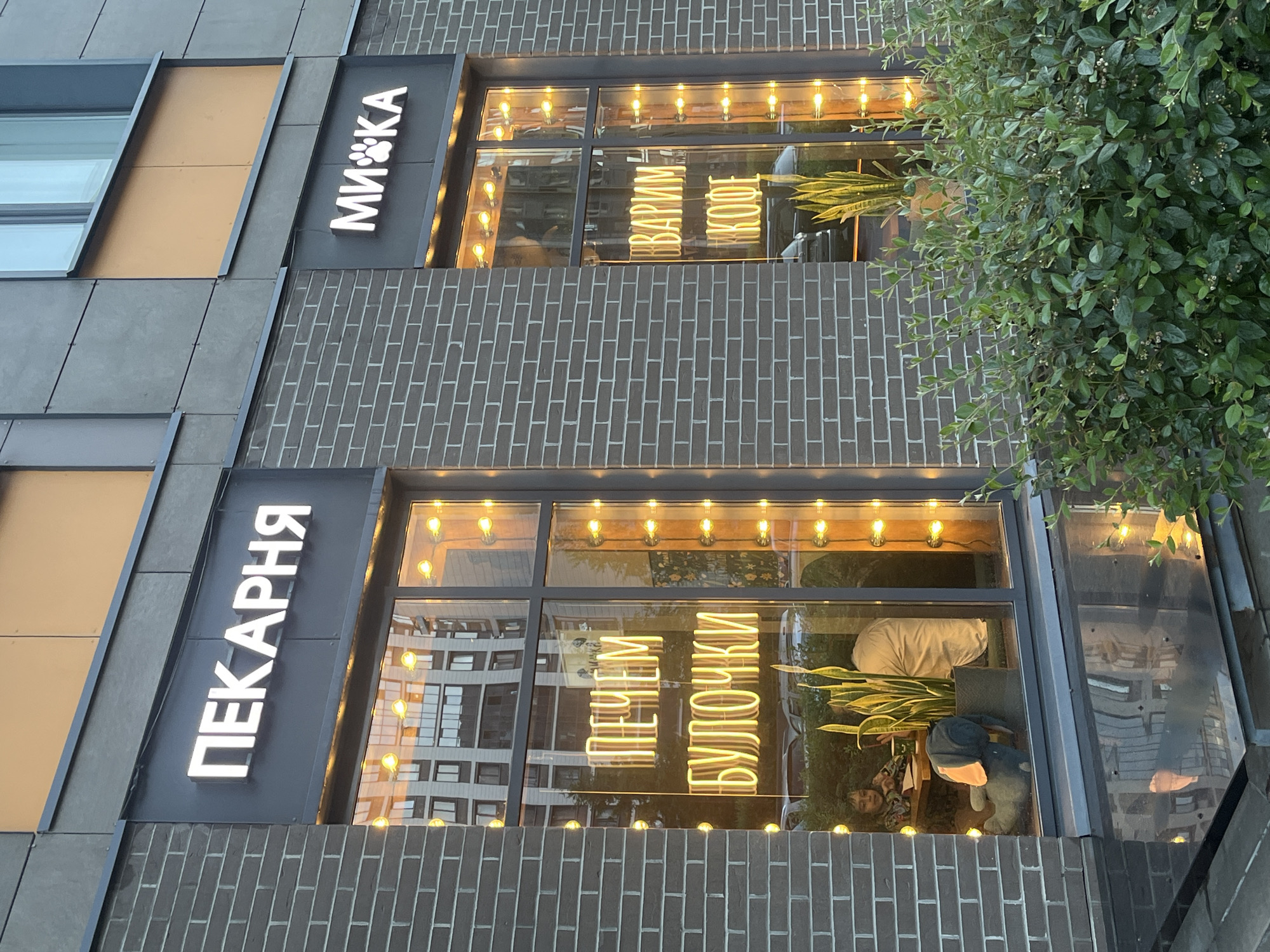 The image shows the exterior of a bakery and a cafe with large windows. The bakery sign, in white illuminated letters, reads "ПЕКАРНЯ" (PEKARNYA), which translates to "Bakery." Below this sign, in bright yellow neon letters within the window, it says "ПЕЧЕМ БУЛОЧКИ" (PECHEM BULOCHKI), meaning "We Bake Buns."
To the right of the bakery, another sign in white illuminated letters reads "МИКА" (MIKA), which includes a paw print graphic. The window of this establishment displays yellow neon text reading "ВАРИМ КОФЕ" (VARIM KOFE), meaning "We Brew Coffee."
Inside the bakery window, a young child sits at a table, facing away from the viewer, with a stuffed animal toy on the floor next to them. A tall potted plant is also visible. The cafe window shows a similar potted plant and reflections of a building.
The buildings are clad in dark brick. The overall lighting suggests it is either early morning or late evening, with artificial lights illuminating the signs and interiors. Green bushes are in the foreground, partially obscuring the lower part of the building. The location is specified as Unknown, Russia.