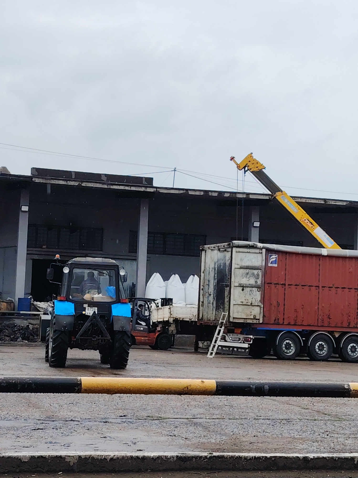 The image depicts an active industrial loading yard in Noviy, Russia, under an overcast sky with light precipitation evident from specks in the air and wet ground. The scene is dominated by heavy machinery involved in material handling operations.
In the foreground, a blue tractor is positioned with its rear facing the viewer. Its license plate, "B 728 YA," is visible. A driver is seated within the tractor's cab. To the right of the tractor, a red forklift is partially visible, likely engaged in moving goods on the ground.
The central activity revolves around a semi-trailer truck carrying two large containers. The front container appears old and weathered, while the rear one is a rusty red. A blue, stylized "F"-like logo is visible on the red container. A white ladder is propped against the truck's side. Extending from behind the truck and arching over the containers is the long, yellow boom of a crane, bearing the "XCMG" logo, indicating ongoing loading or unloading.
Behind the forklift and to the left of the containers, several large white bulk bags, presumably containing industrial materials like fertilizer or grain, are stacked near a building. The background features a large, dark-colored industrial building or warehouse with multiple loading bay entrances, suggesting a busy logistics hub. The overall impression is one of a functional outdoor industrial facility focused on transport and material management.