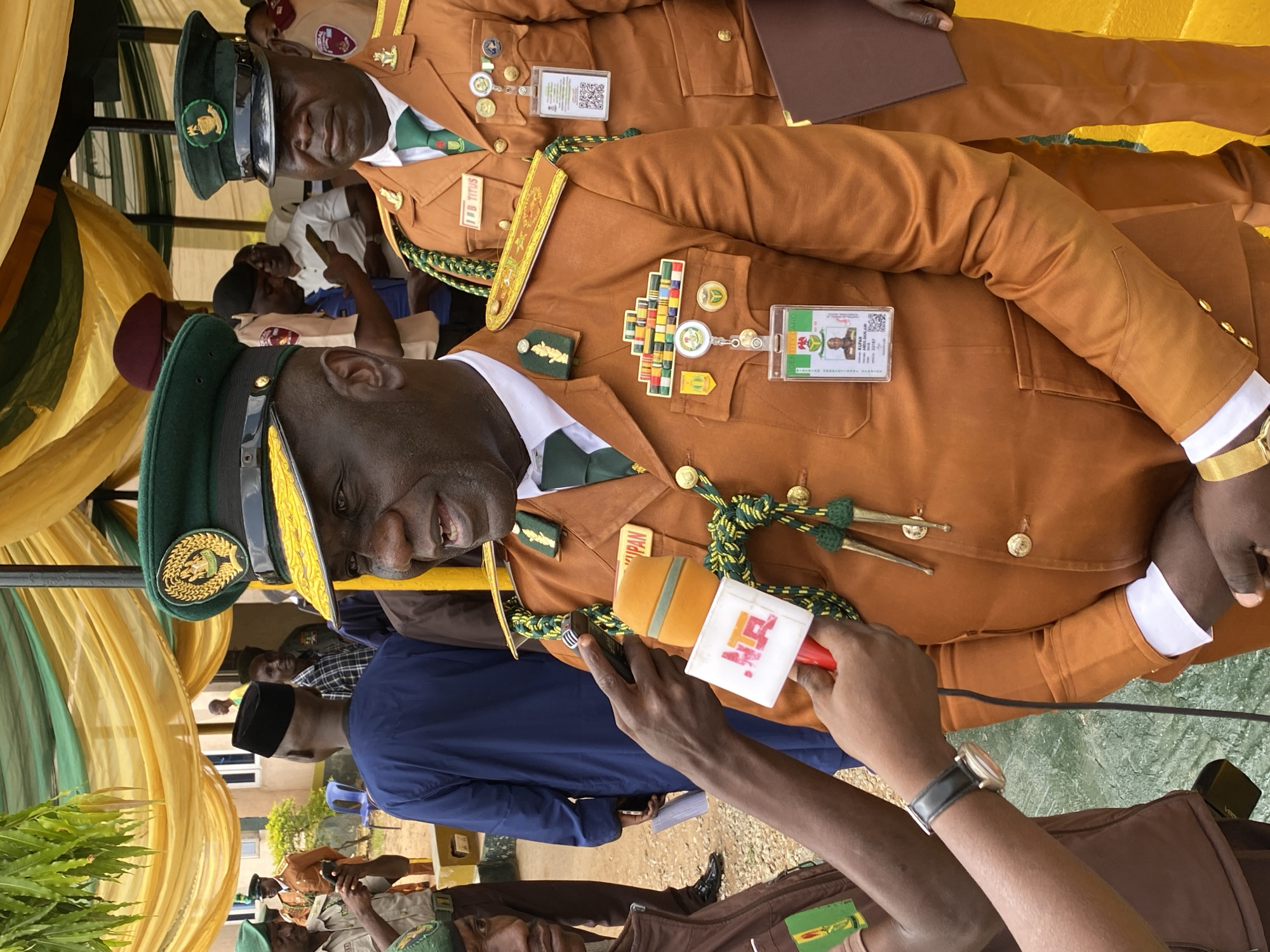The image depicts an outdoor event, likely a ceremony or official gathering, set in Kaduna, Nigeria. The primary focus is on a group of men in formal, matching brown uniforms with green accents and dark green peaked caps. The uniforms are adorned with various medals, badges, and insignia, suggesting a formal occasion.
One man in the foreground, facing the camera, is being interviewed. He is wearing a brown uniform with a white shirt and a green tie. He has a microphone with the "NTA" logo (Nigerian Television Authority) held up to him, indicating he is a subject of a broadcast or interview. His expression appears animated and engaged, suggesting he is speaking or listening intently. Another man, slightly behind and to the left, also in uniform, is partially visible, looking towards the interviewee.
Behind these central figures, other individuals in similar uniforms are visible, some participating in the event, others observing. The setting includes draped yellow and green fabrics, possibly forming a backdrop or canopy, and hints of greenery and buildings in the background. The lighting suggests it is daytime.
Visible text includes the "NTA" logo on the microphone and an identification card attached to the interviewee's uniform which reads "AB TITUS" and appears to have more text that is illegible. Another identification card is visible on a person to the right, reading "KUPAK" and "2023-08-14" and "23:17:57".
