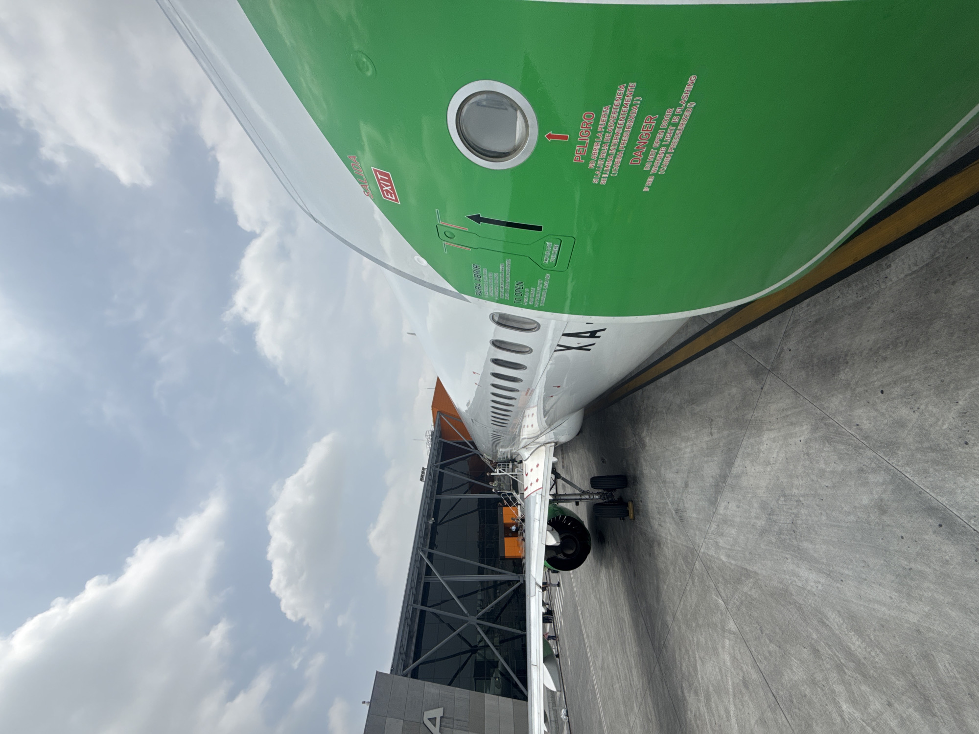 An airplane is prominently featured, parked at an airport gate, viewed from a low angle on the tarmac. The aircraft's fuselage is white with a distinctive bright green upper section and an orange tail fin, though only a small part of the tail is visible. A major focus is on an emergency exit door on the green section of the fuselage, which has a small porthole window.
The door features red text warnings and instructions in both Spanish and English. Visible text includes "SALIDA," "EXIT," and an arrow pointing upwards. Below this, there are instructions "PARA ABRIR" and "TO OPEN" with an illustration showing how to operate the door. Further right, "PELIGRO" and "DANGER" are printed, followed by a warning: "NO ABRIR LA PUERTA SI LA LUZ ROJA DE ADVERTENCIA SE ILUMINA INTERMITENTEMENTE (CABINA PRESURIZADA!)" and its English translation: "DO NOT OPEN DOOR IF RED WARNING LIGHT IS FLASHING (CABIN PRESSURIZED)." The registration "XA" is also visible on the white part of the fuselage.
The airplane's landing gear and part of an engine are visible, alongside a modern-looking terminal building with large glass panels and a connecting jet bridge structure. The ground is a concrete tarmac with yellow lines. The sky above is cloudy, suggesting a daytime scene. The location is Apodaca, Mexico. No people are visible.