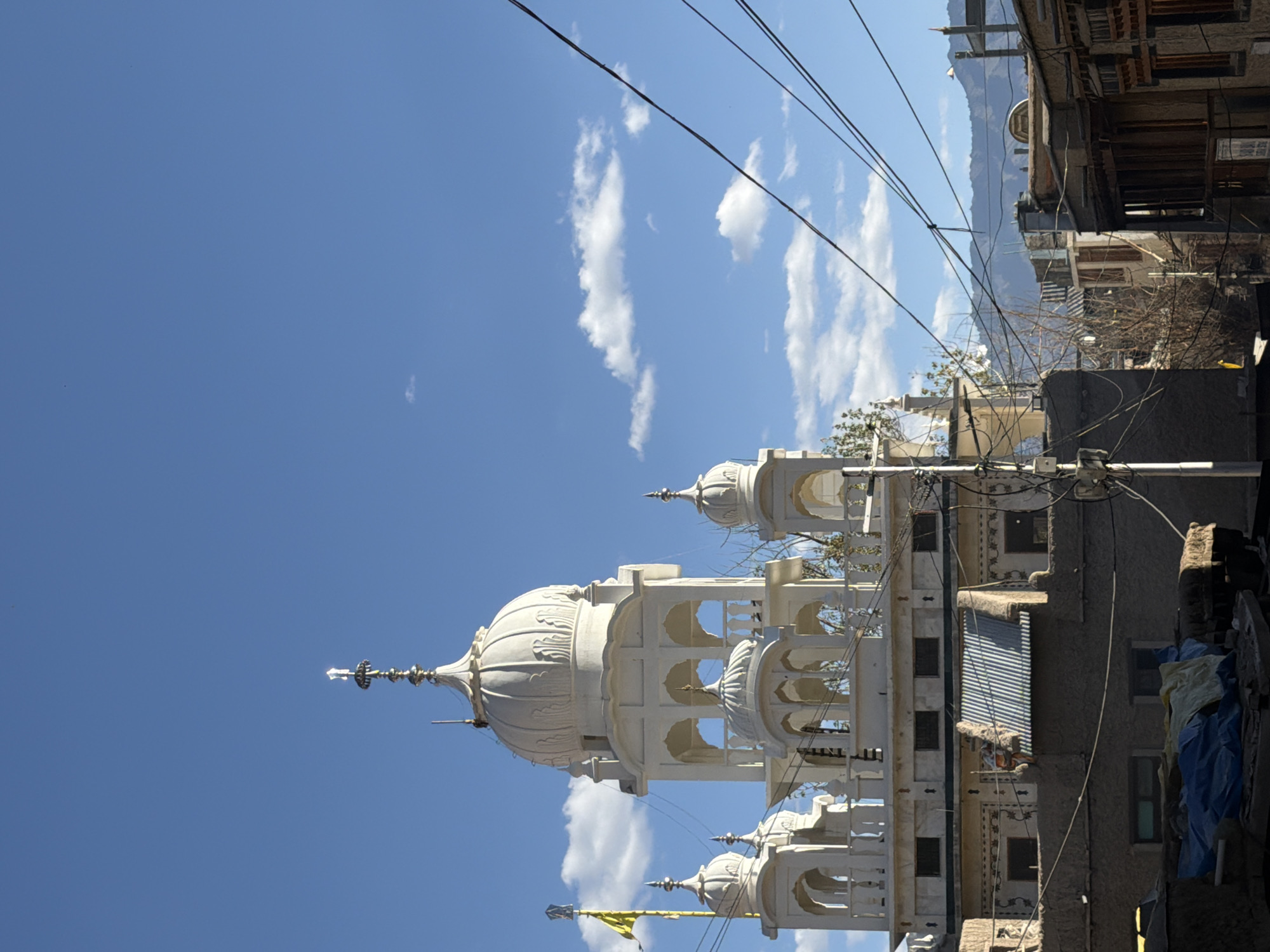 The image captures a Gurudwara in Leh, India, under a bright blue sky dotted with scattered clouds. The Gurudwara is a white, multi-tiered structure with several domes, including a prominent central dome topped with a finial. Intricate architectural details are visible, such as arched openings and decorative railings. A yellow flag flutters from a flagpole to the left of the Gurudwara.
In the foreground, there are several layers of buildings with a more rustic, earthy appearance. These structures feature corrugated metal roofs and windows. Visible are numerous power lines crisscrossing the frame, contributing to a sense of urban or semi-urban development. In the distance, mountains form a backdrop, suggesting the mountainous terrain of Leh. The overall impression is one of a culturally significant religious site situated within a lived-in environment. No people are clearly identifiable in the scene.