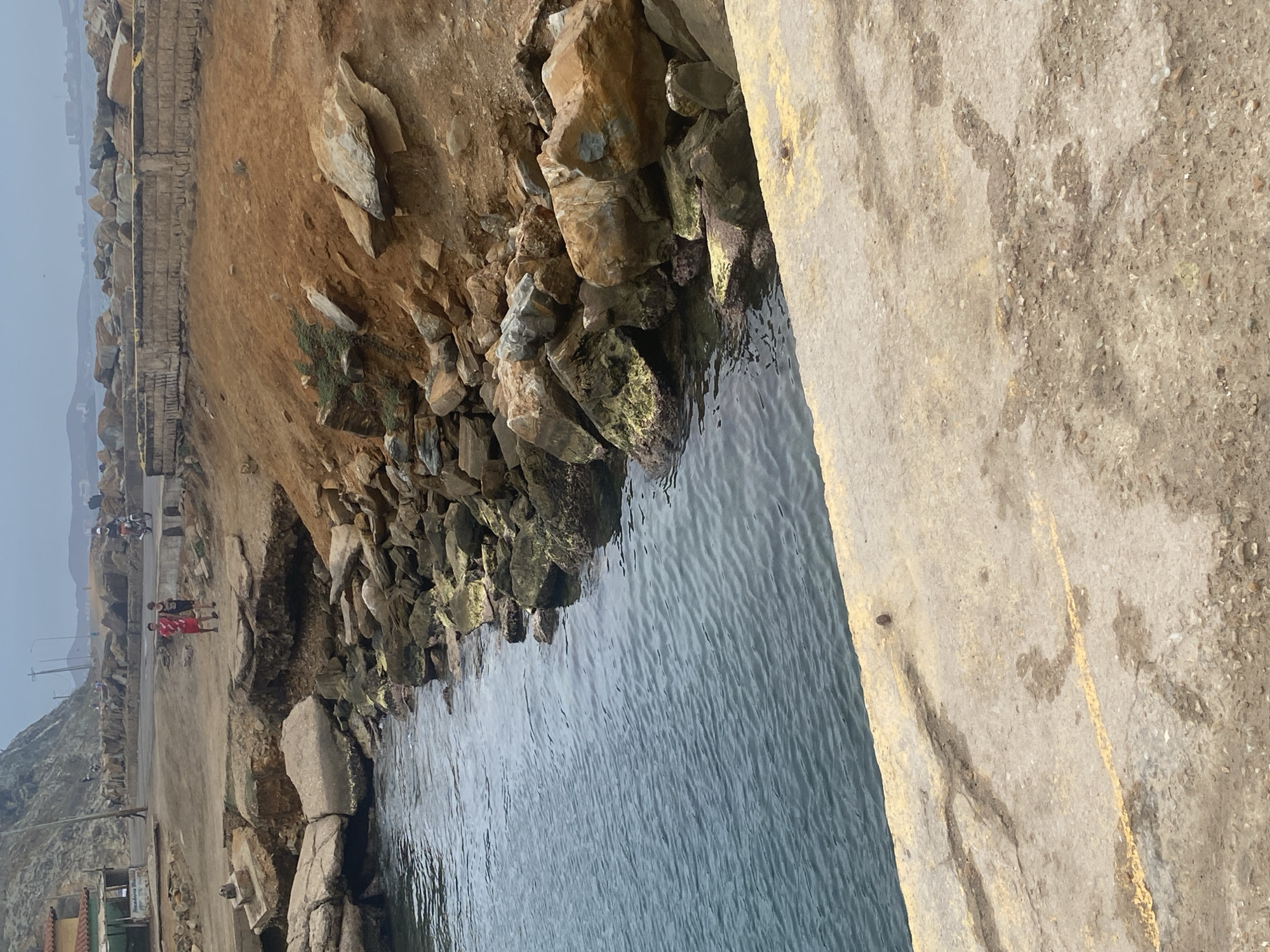 The image shows a shoreline with a mix of rocky cliffs, sandy terrain, and a concrete structure abutting the water.
People are visible in the distance on a path running alongside the cliff. The concrete structure seems to be a pier or a breakwater, and the water is relatively calm. The sky appears overcast, suggesting a cloudy or hazy day. The image's location is likely along the coast of Porlamar, Venezuela.