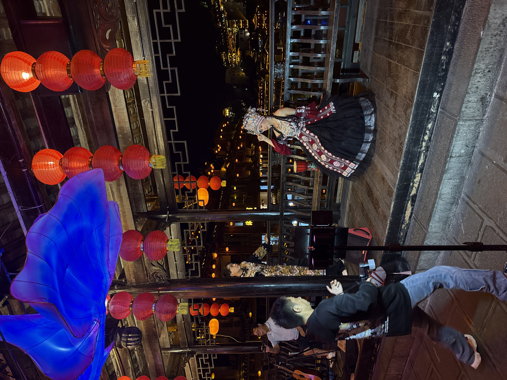 The image captures an outdoor scene at night in Furong, China, likely within a historic or tourist district. A performer dressed in a traditional embroidered black dress with ornate headwear poses on an elevated walkway. Behind her is a railing with a view of the cityscape, illuminated with lights. Below, two performers and another person are observed: one performer is wearing a patterned garment. Further down, a man in a black top and blue pants, possibly a photographer or onlooker, is present, along with another man in a white shirt. Strings of red lanterns adorn the walkway. A large blue installation is on the bottom left corner. The setting indicates a cultural event or performance in a tourist location within Furong.