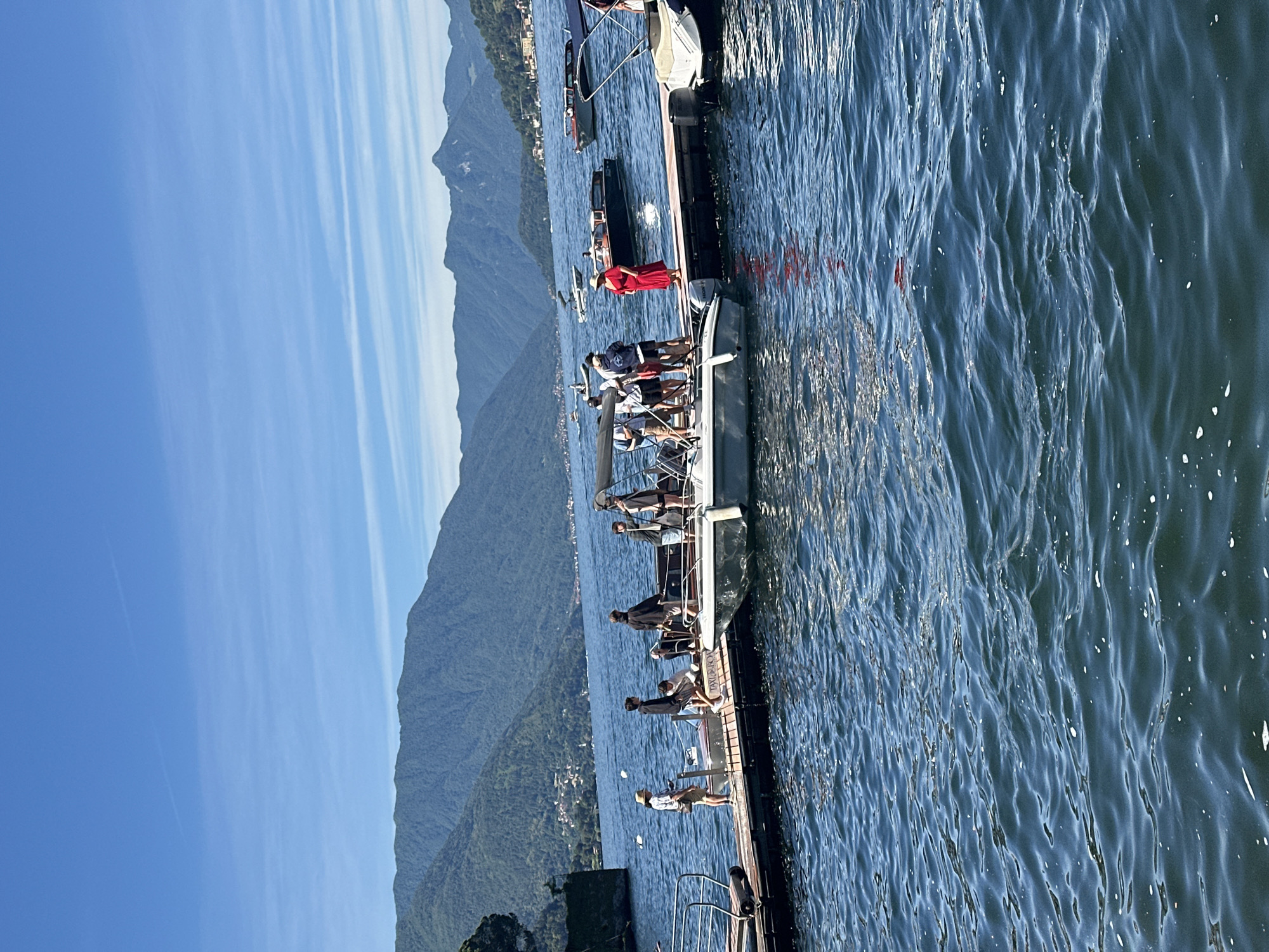 In Varenna, Italy, a bright, sunny day is depicted on a lake with mountainous terrain in the background. Several people are gathered on a wooden dock, some standing, others sitting on the edge. A few small boats are docked alongside the pier, and one is visible further out on the water. One person, wearing a red dress, stands out on the dock. The water is a deep blue with gentle ripples, and the sky is a clear, vibrant blue with a few wispy clouds.
