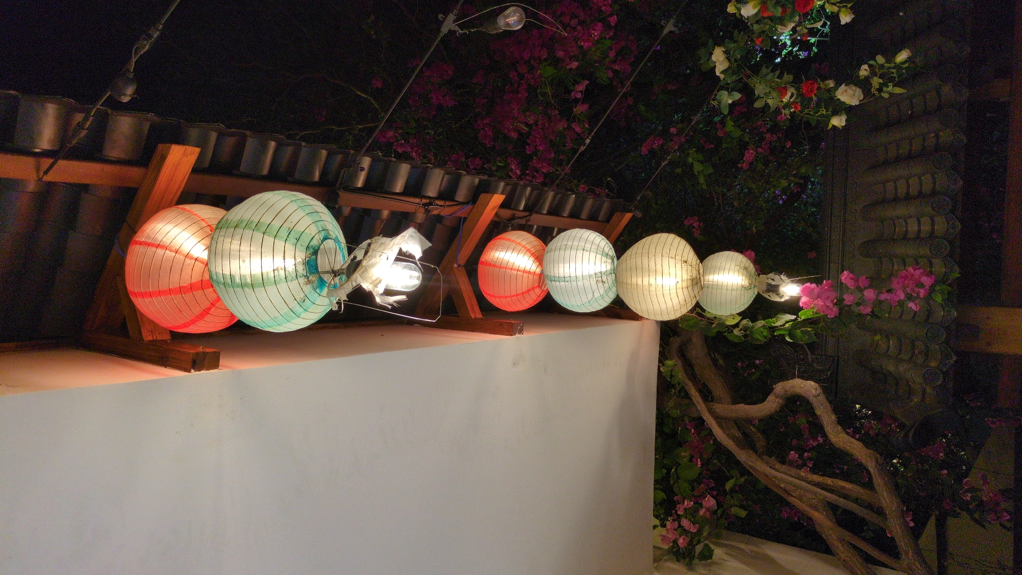 The image shows a row of illuminated paper lanterns hung along a low, white wall. The lanterns are multicolored, with hues of red, green, and white. They are suspended by a string of lights that is attached to a wooden frame covered by what appears to be a corrugated metal roof. Behind the lanterns, bougainvillea flowers and other plants grow up a wall or structure. The scene appears to be outdoors at night. This photo may have been taken in the city of Phan Rang – Tháp Chàm, Vietnam.