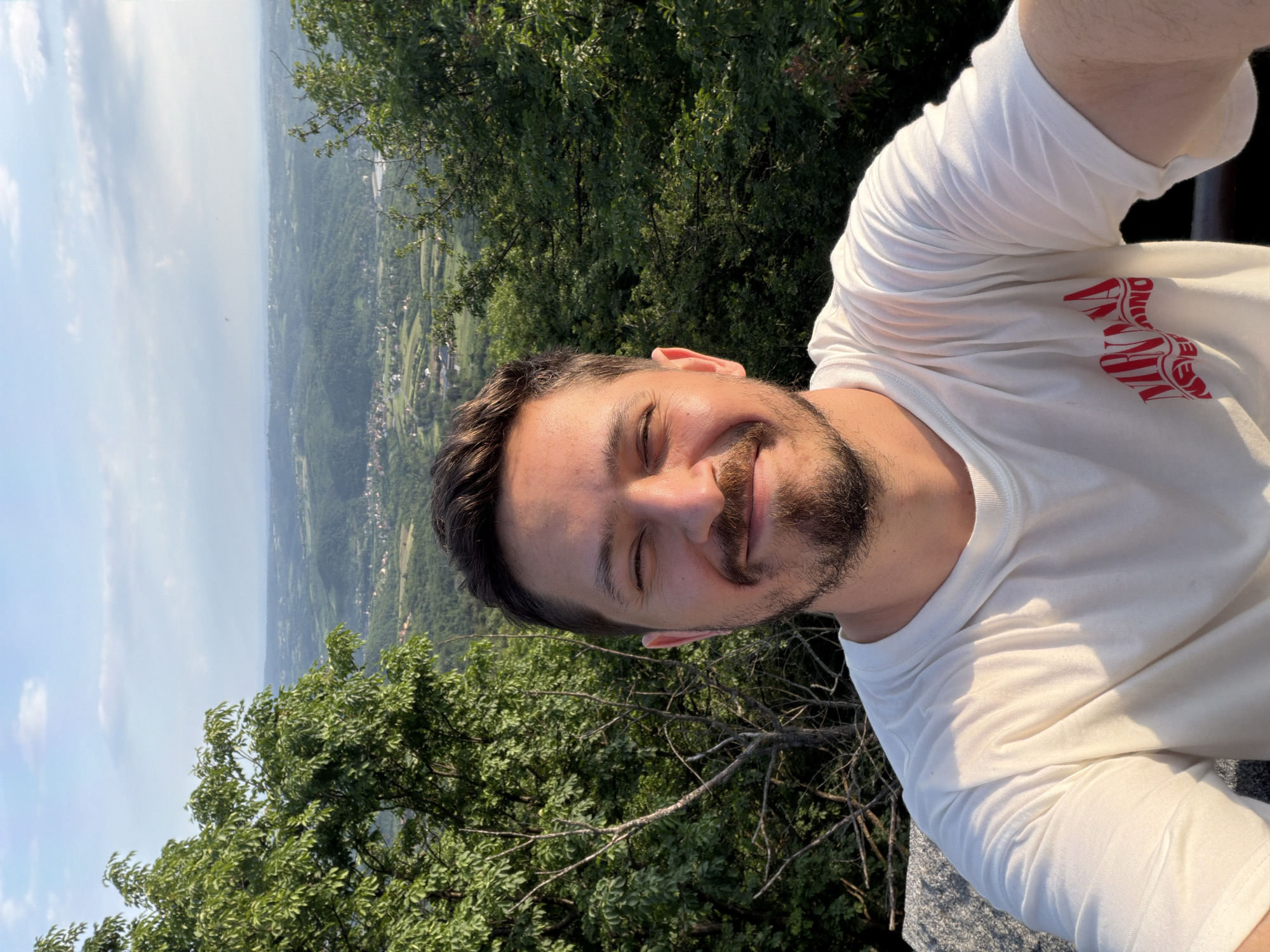 A selfie depicts a man with his eyes closed, a slight smile, and a beard. He wears a white t-shirt with a red graphic. Behind him, there is dense green foliage and a panoramic view of a landscape under a partly cloudy sky. Based on the landscape and surrounding environment, it is possible that this photo was taken in the municipality of Vozdovac, Serbia.