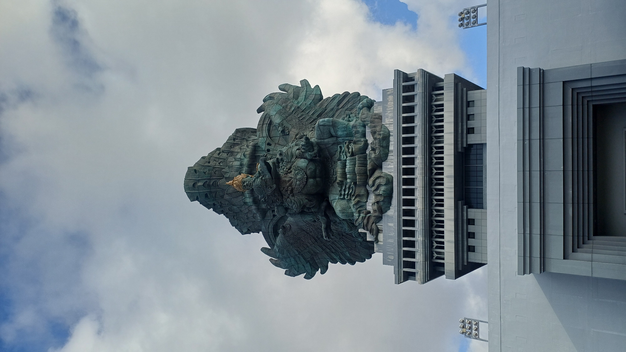 The image depicts the Garuda Wisnu Kencana statue in Kuta Selatan, Indonesia. The statue, primarily composed of bronze-green metal, features Garuda carrying Vishnu. The statue is positioned adjacent to a grey building with a geometric, stepped design. The background reveals a cloudy sky with patches of blue. The image is oriented on its side.