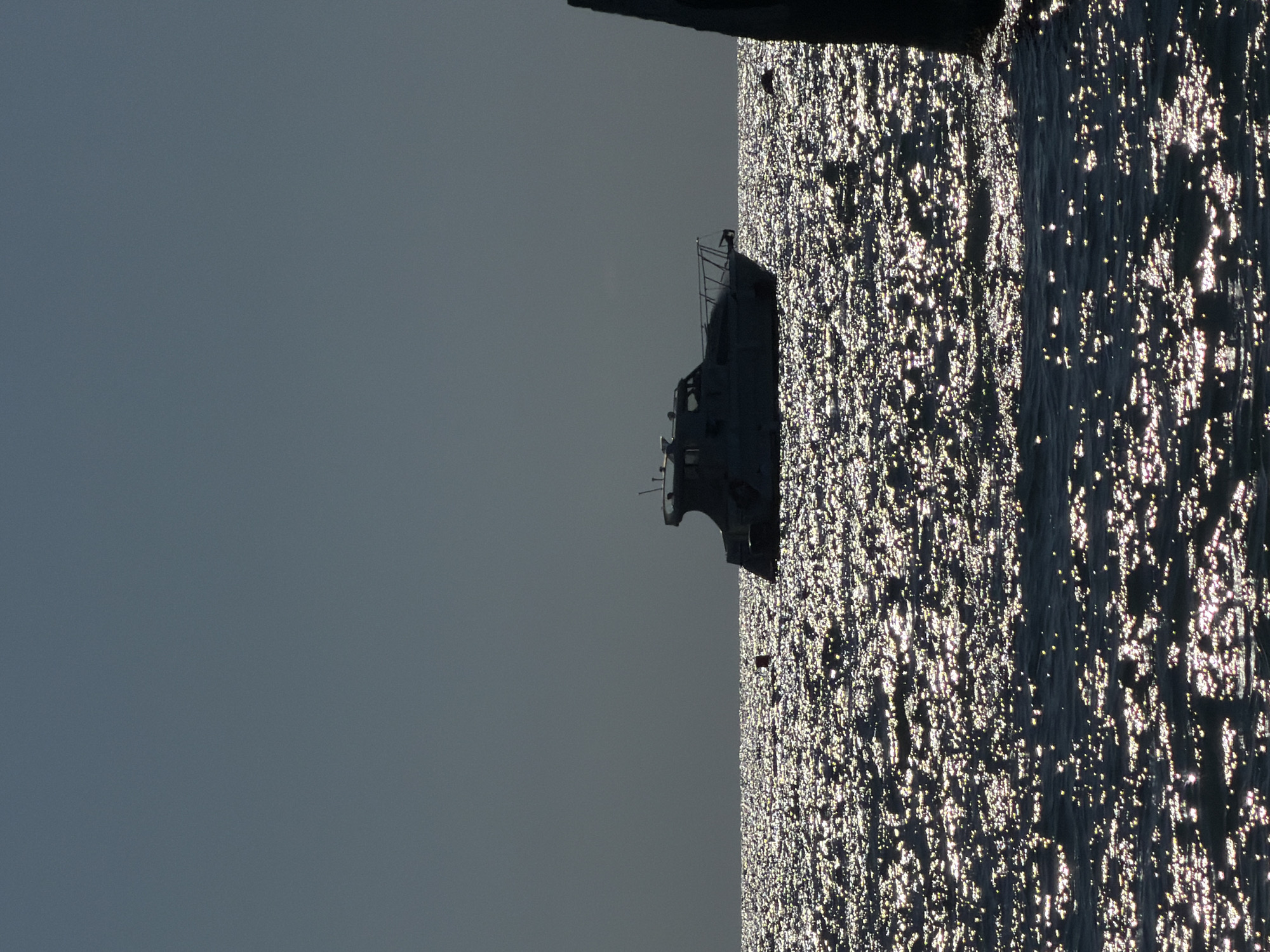 A silhouette of a motorboat is visible on the shimmering surface of the sea. The water is aglow with reflected sunlight, creating a glittering effect. The sky above is a muted, dark gray. On the right side of the frame, a dark, vertical structure, possibly a pier or a building element, juts into the scene from the edge. A small red buoy is also present in the water, to the left of the boat. The image appears to capture a serene moment at sea, likely during dawn or dusk given the lighting conditions and the silhouette effect. The location is specified as the city of Sochi, Russia.