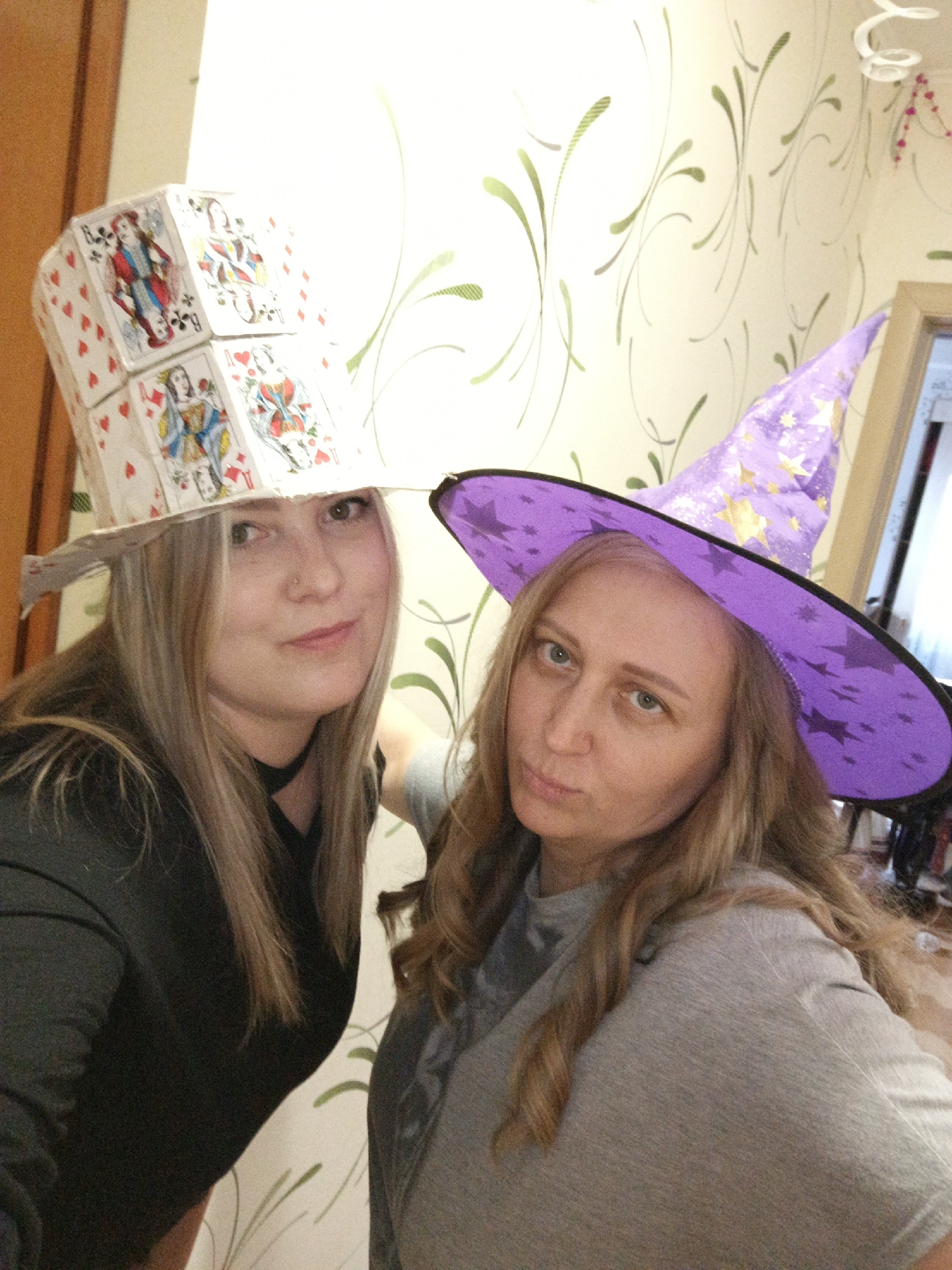 Two women are posing indoors, wearing hats. The woman on the left has long blonde hair and is wearing a black top and a white hat made of playing cards. The woman on the right has wavy brown hair and is wearing a gray top and a purple witch hat with gold stars. The background is a wall with a light-colored floral pattern. The location is Марковский, Russia.