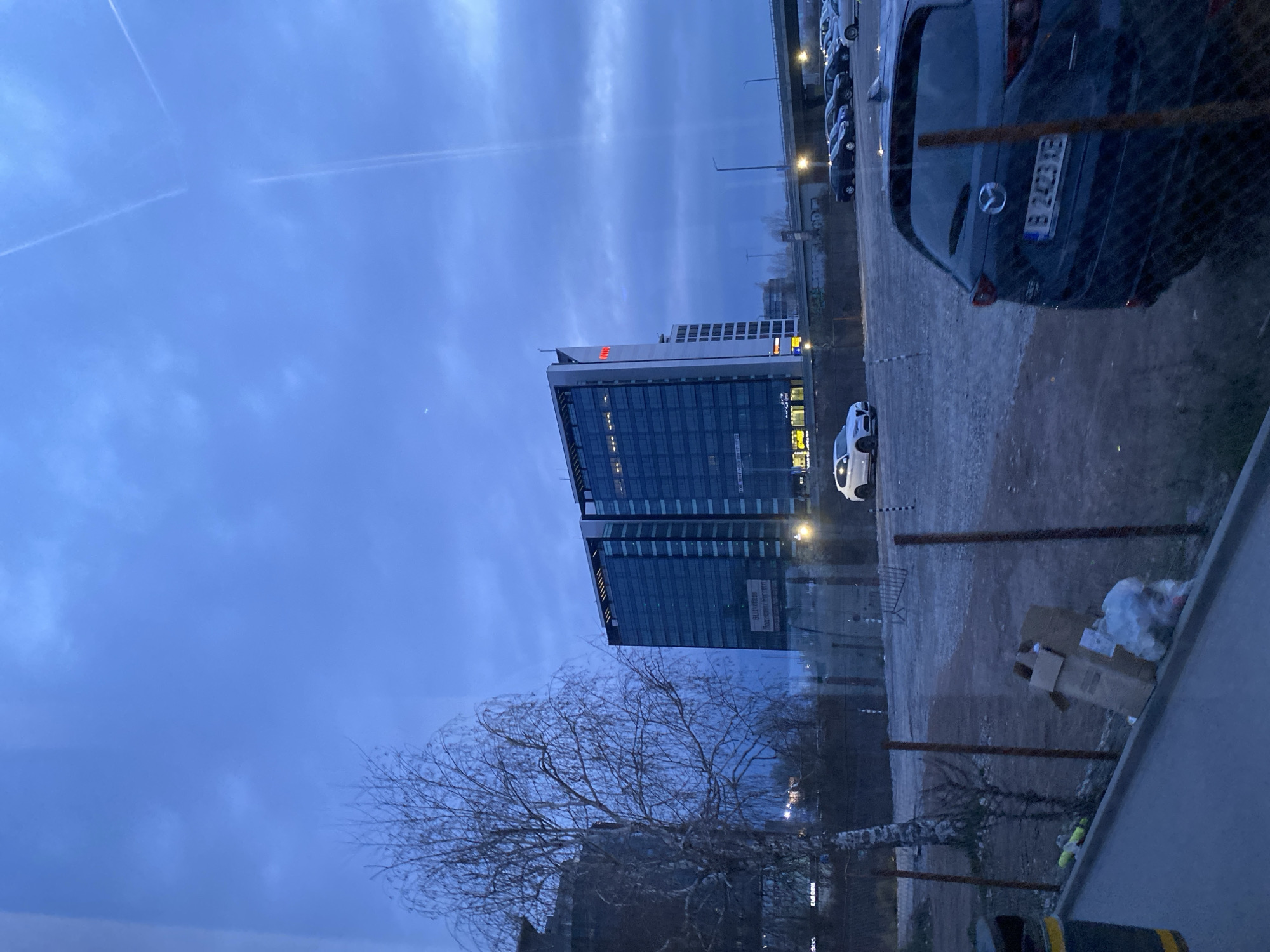 The image shows an exterior view of a building and surrounding area. The prominent structure is a modern high-rise building with a glass facade. The building displays an "A" logo with a red top and white base. A Mazda car is parked with license plate "B 2423 XE". There is a row of parked cars in the background, a tree, and a sky with clouds and vapor trails. The presence of the license plate suggests that this location is in Sofia, Bulgaria. There is litter in the foreground.