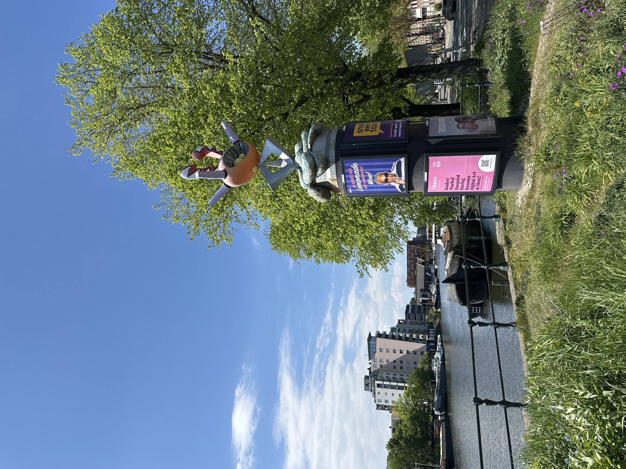 The image depicts an outdoor scene in Amsterdam, Netherlands. A canal is visible, lined by buildings and vegetation. A prominent advertising column stands alongside the canal, featuring advertisements for lemonade, a taxi service, and the MOCO Museum, highlighting artists like Banksy, Warhol, and Kusama. On top of the advertising column is a peculiar sculpture of an orange figure with outstretched arms. A large tree provides a backdrop, with a blue sky visible above.
