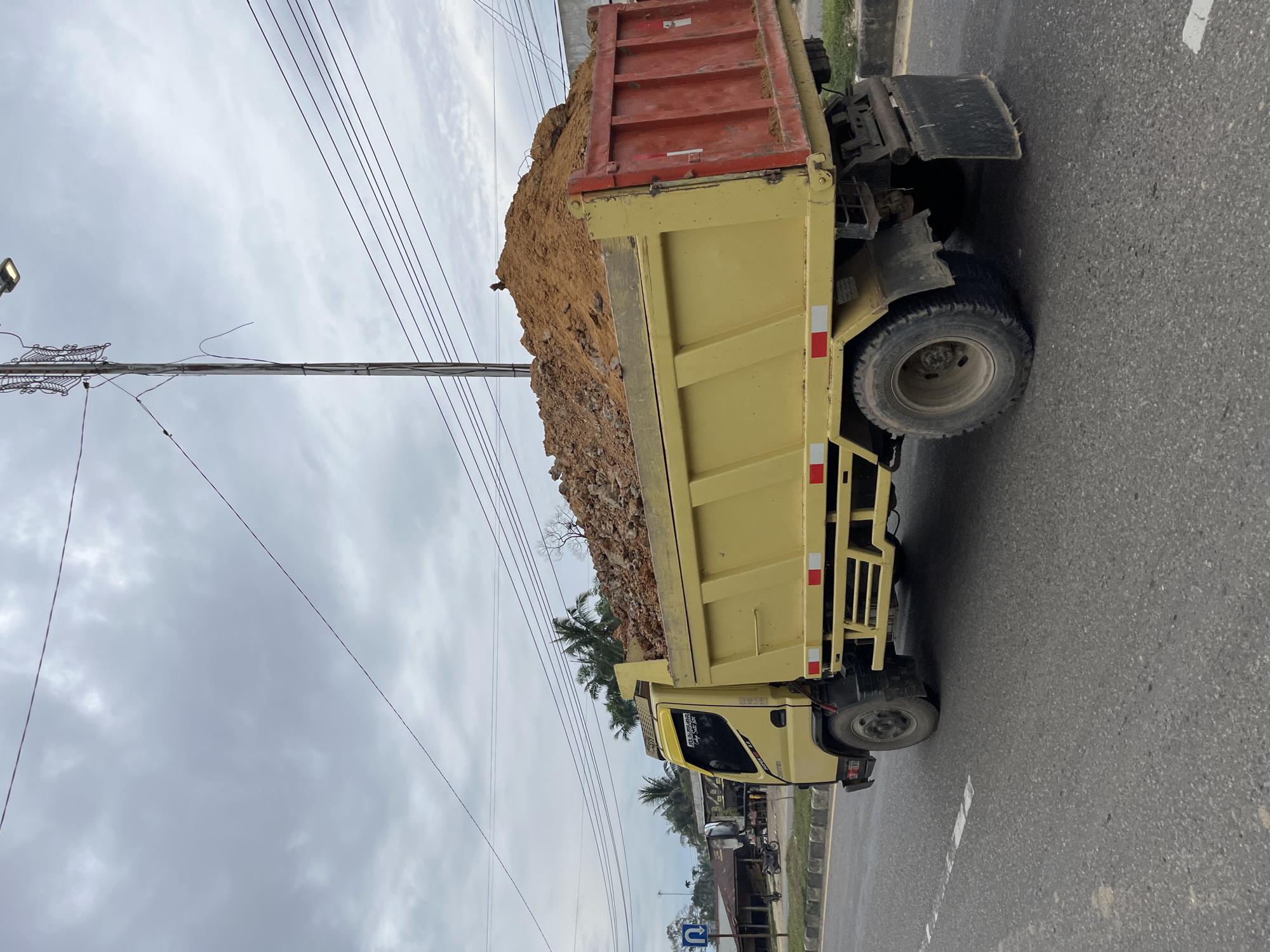 In the city of Tanahterban, Indonesia, a yellow dump truck is captured in motion on an asphalt road under an overcast sky. The truck is laden with a large pile of soil and rocks, which fills its bed and spills slightly over the sides. The truck's side is visible, showing red accents on the rear, and reflective safety strips on the yellow body. In the background, there are buildings, palm trees, a utility pole with wires, and a street sign indicating a U-turn is permitted. The overall scene suggests ongoing construction or land movement activities in the area.