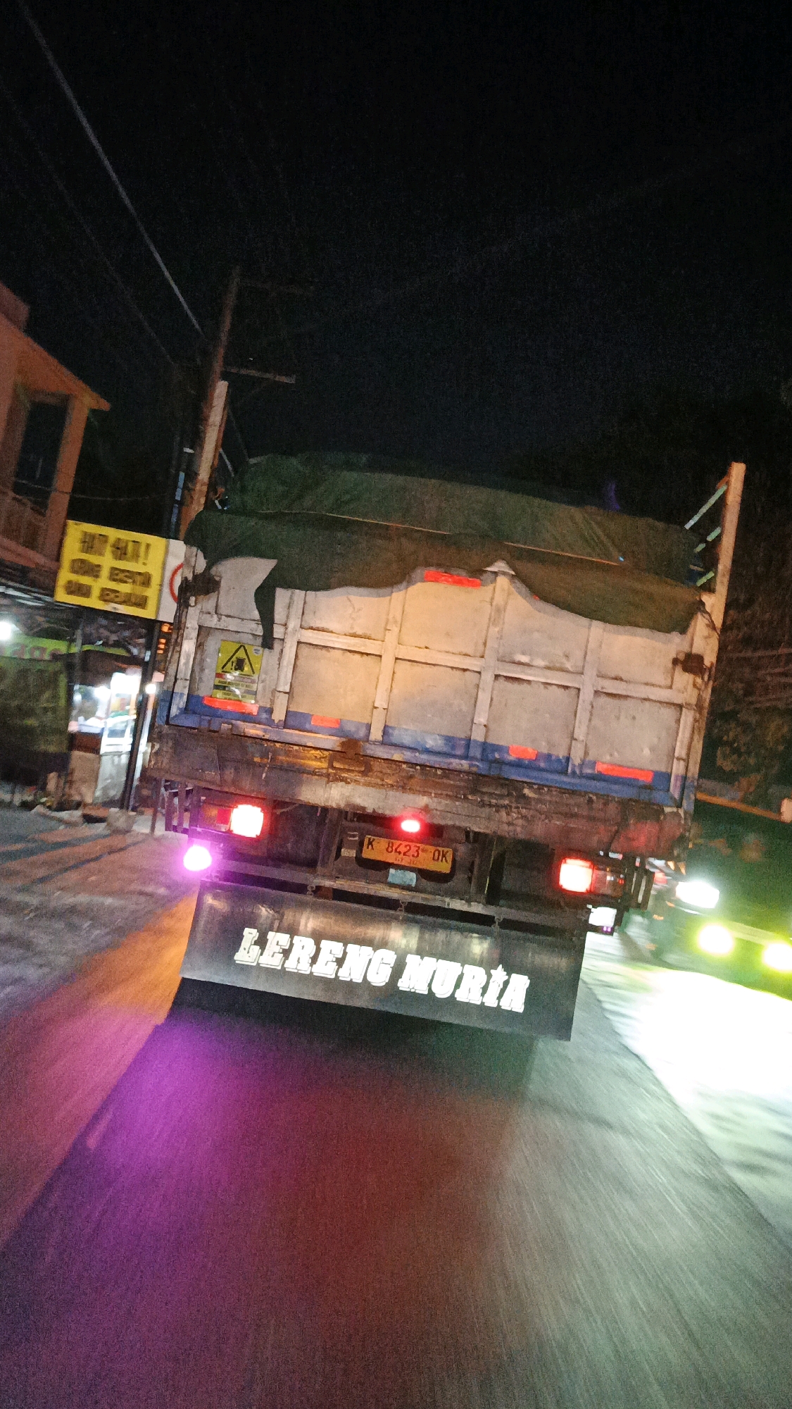 The image depicts the rear of a dump truck driving at night on a street in Surakarta, Indonesia. The truck's license plate reads "K 8423 OK". The truck is labeled "LERENG MURIA" on its tailgate. The street is wet, suggesting recent rain or a wet road surface, and is illuminated by the truck's taillights and the headlights of other vehicles in the background, creating reflections and streaks of light. There are also overhead power lines and streetlights visible. A building with signage is partially visible on the left side of the image. The overall impression is a typical nighttime scene on an urban road in Indonesia.