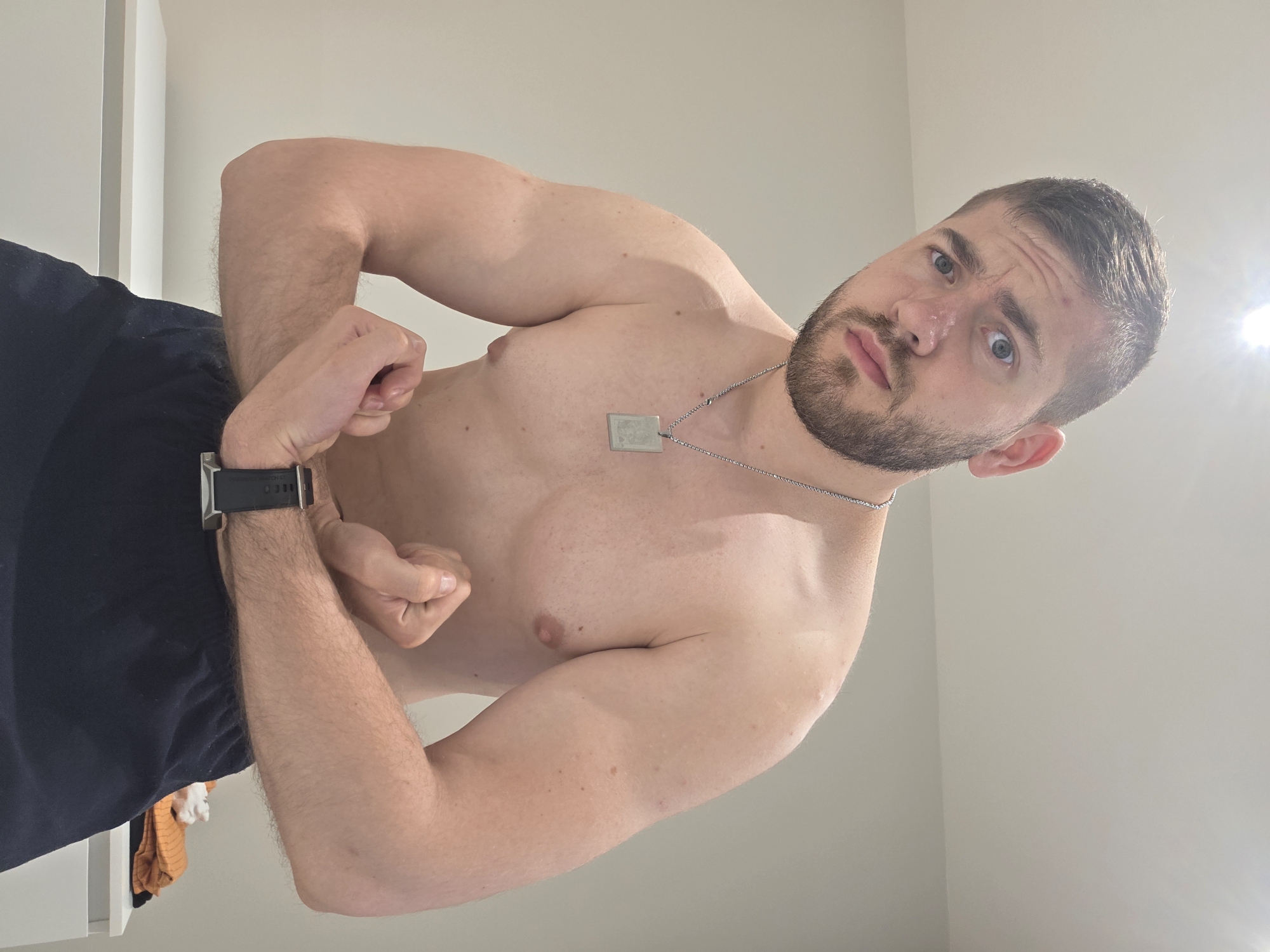 A shirtless man with a beard is flexing his biceps. He is wearing dark shorts and a watch on his left wrist. He also has a silver chain with a dog tag around his neck. The background is a plain wall. The location is not discernable from the image.