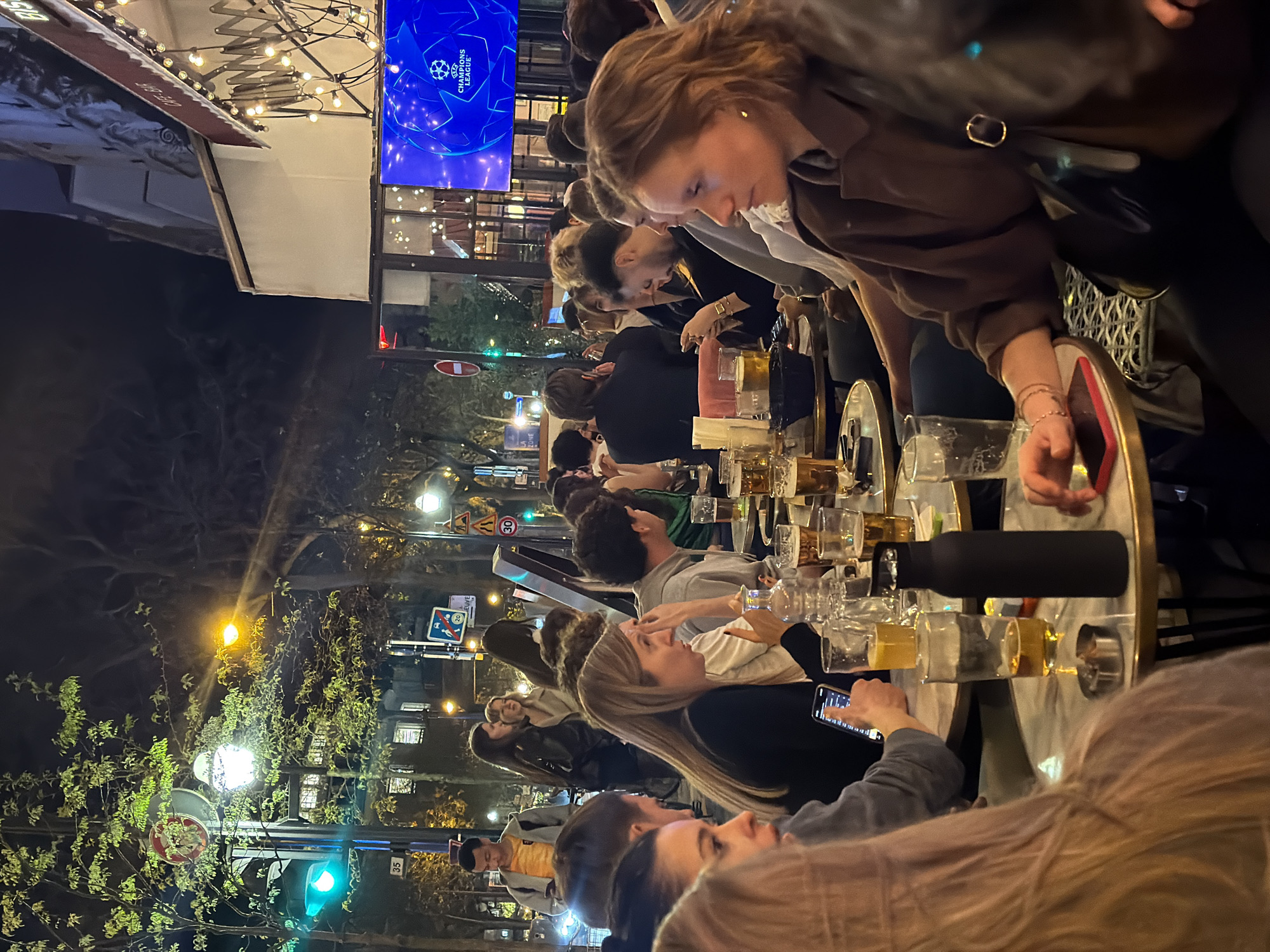 A night scene depicts an outdoor restaurant in Paris, France, where people are gathered at tables filled with drinks. The ambiance is illuminated by overhead string lights and street lamps, creating a warm atmosphere. Patrons are engaged in conversation and using their mobile phones. A large screen displays the Champions League logo, suggesting a sporting event is being shown. Street signs and trees line the background, giving context to the Parisian setting.