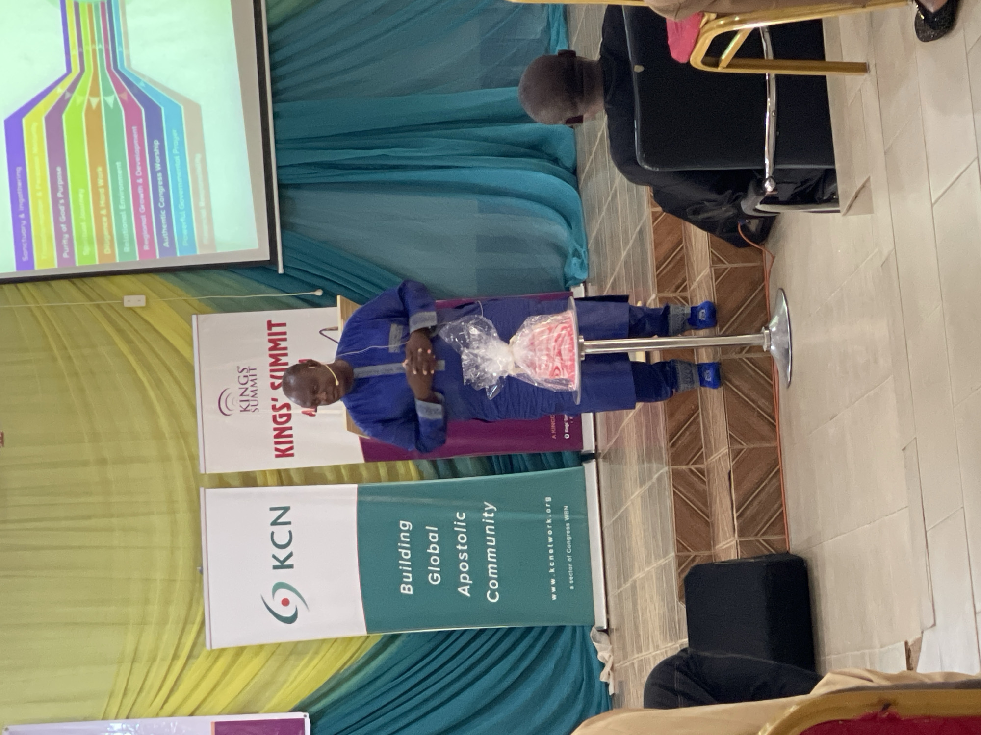 The image shows a man standing at a lectern with two pull-up banners behind him that say "KCN Building Global Apostolic Community" and "Kings Summit". The man is wearing a blue suit. He is holding a package wrapped in clear plastic. Above him, a projection screen displays text including "Sanctuary & Ingathering", "Purity of God's Purpose", "Relational Environment", and "Authentic Congress Worship". The setting appears to be indoors, likely a conference or event in Akwanga, Nigeria.
